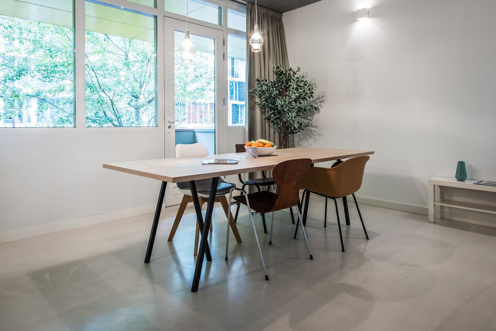 Eetkamer met een houten tafel, stoelen en grote ramen met uitzicht op bomen.