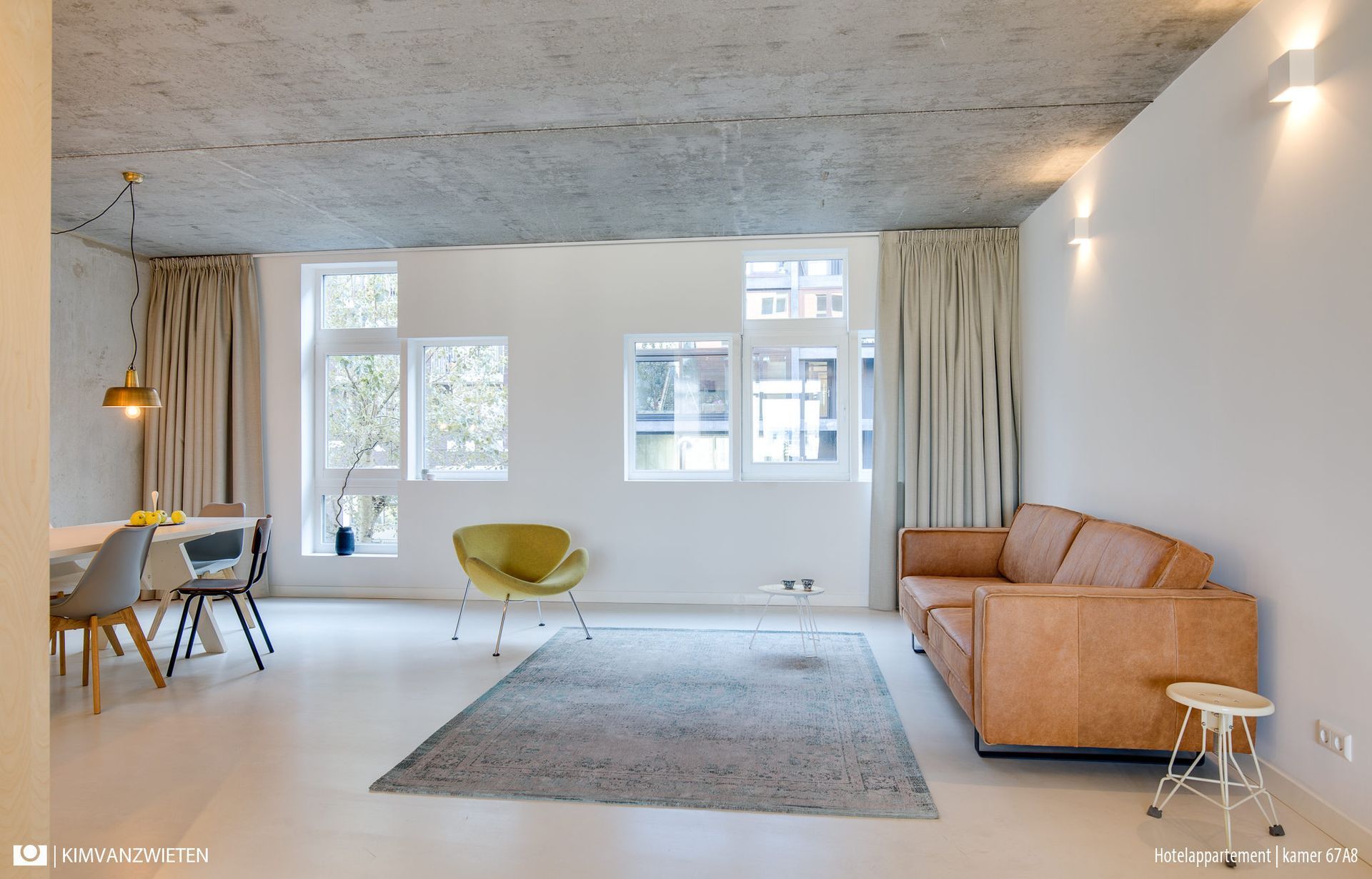 Open woonruimte met een bank, vloerkleed, eettafel en ramen. Witte muren en plafond.