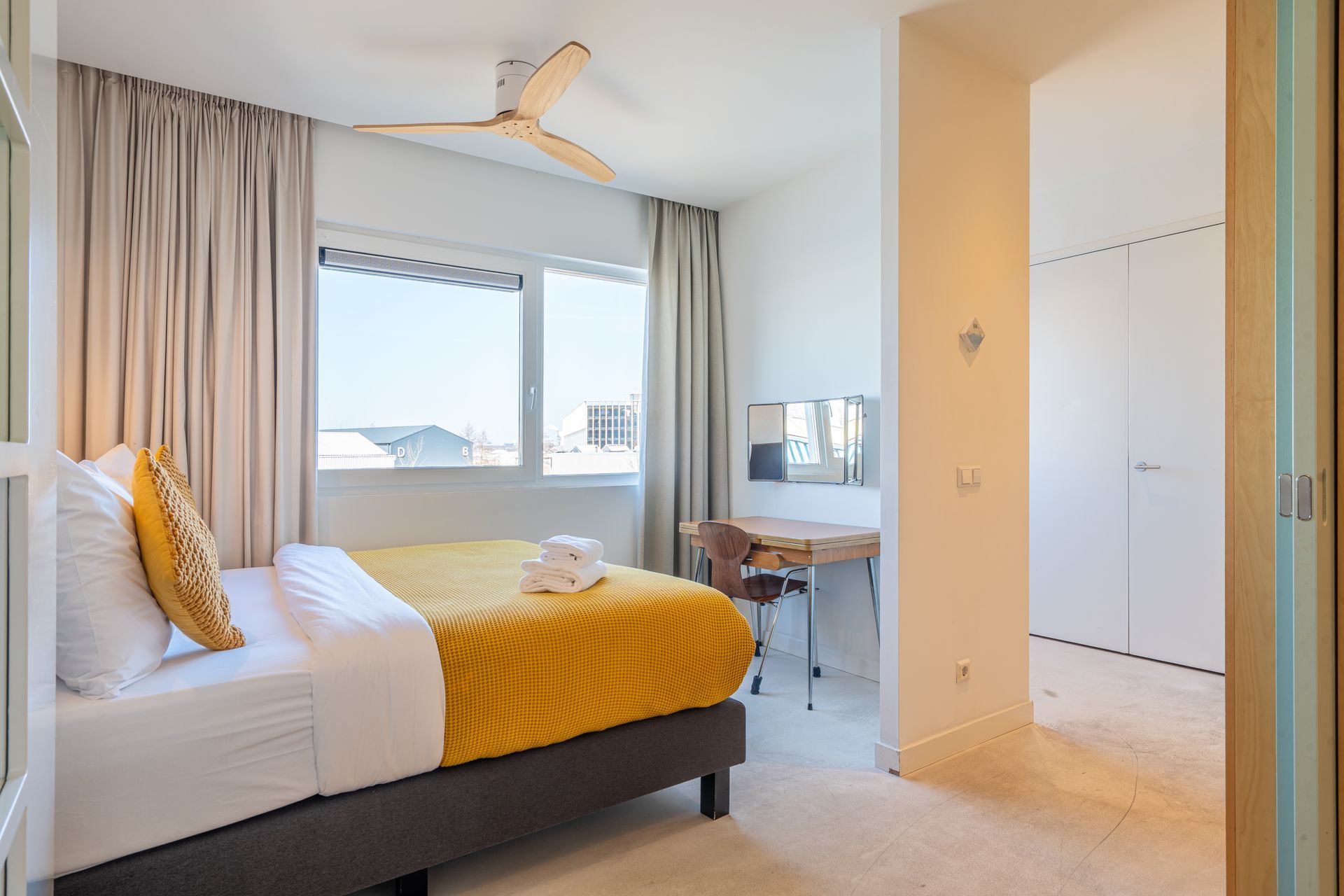 Een moderne, lichte slaapkamer met een bed met een geel dekbed, een bureau, een raam met gordijnen en een plafondventilator.