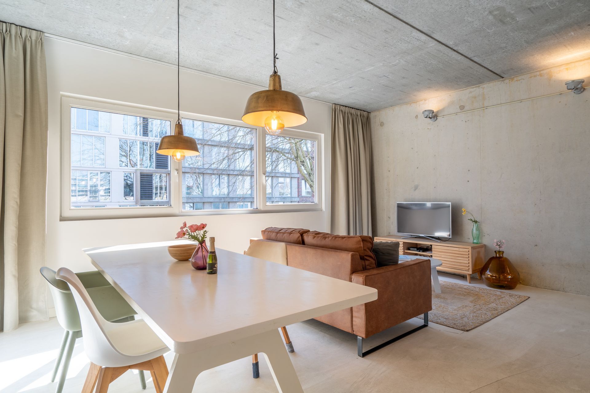 Een licht, modern appartement met een eethoek op de voorgrond en een woonkamer met een leren bank op de achtergrond.