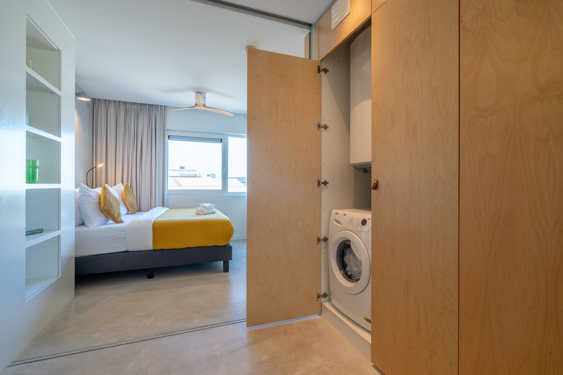 Een hotelkamer met een bed met geel beddengoed, gezien vanuit een aangrenzende ruimte met een wasmachine in een houten kast.