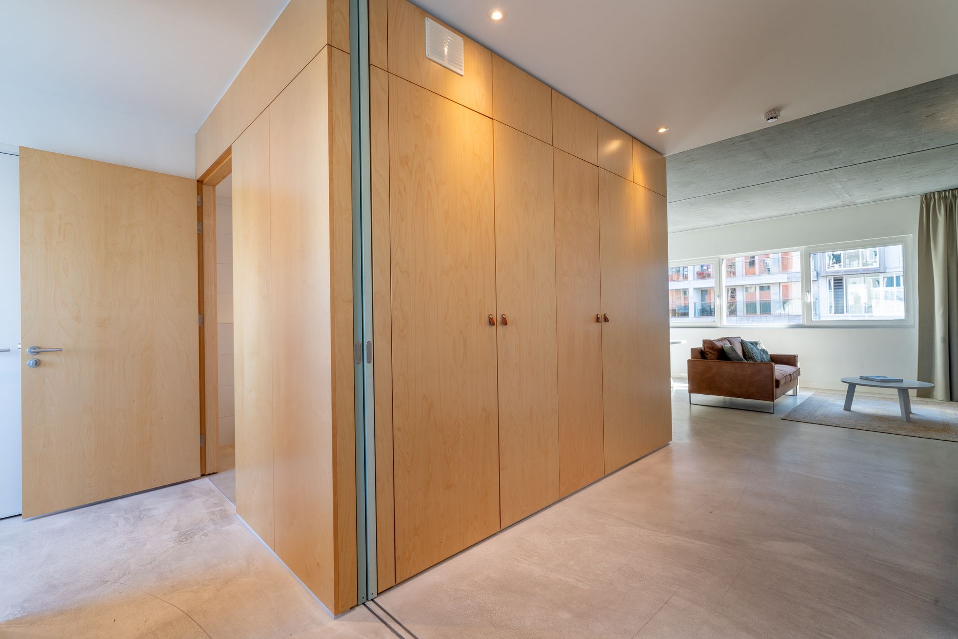 Een scheidingswand van licht hout scheidt de entree van een lichte woonkamer met een bruine bank.