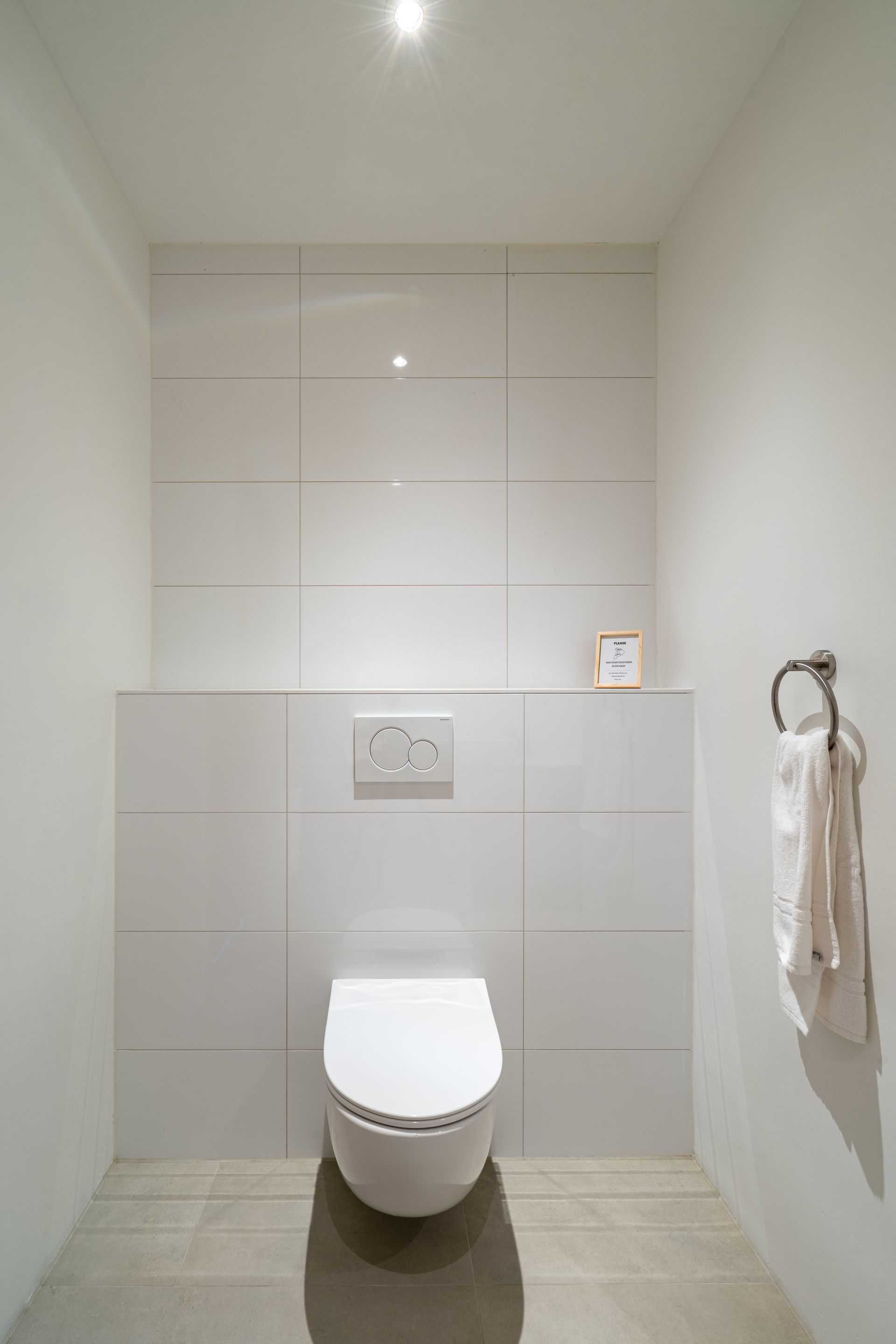 Een moderne, minimalistische badkamer met een wit wandtoilet, lichtgekleurde tegelwanden en een handdoekring aan de zijkant.