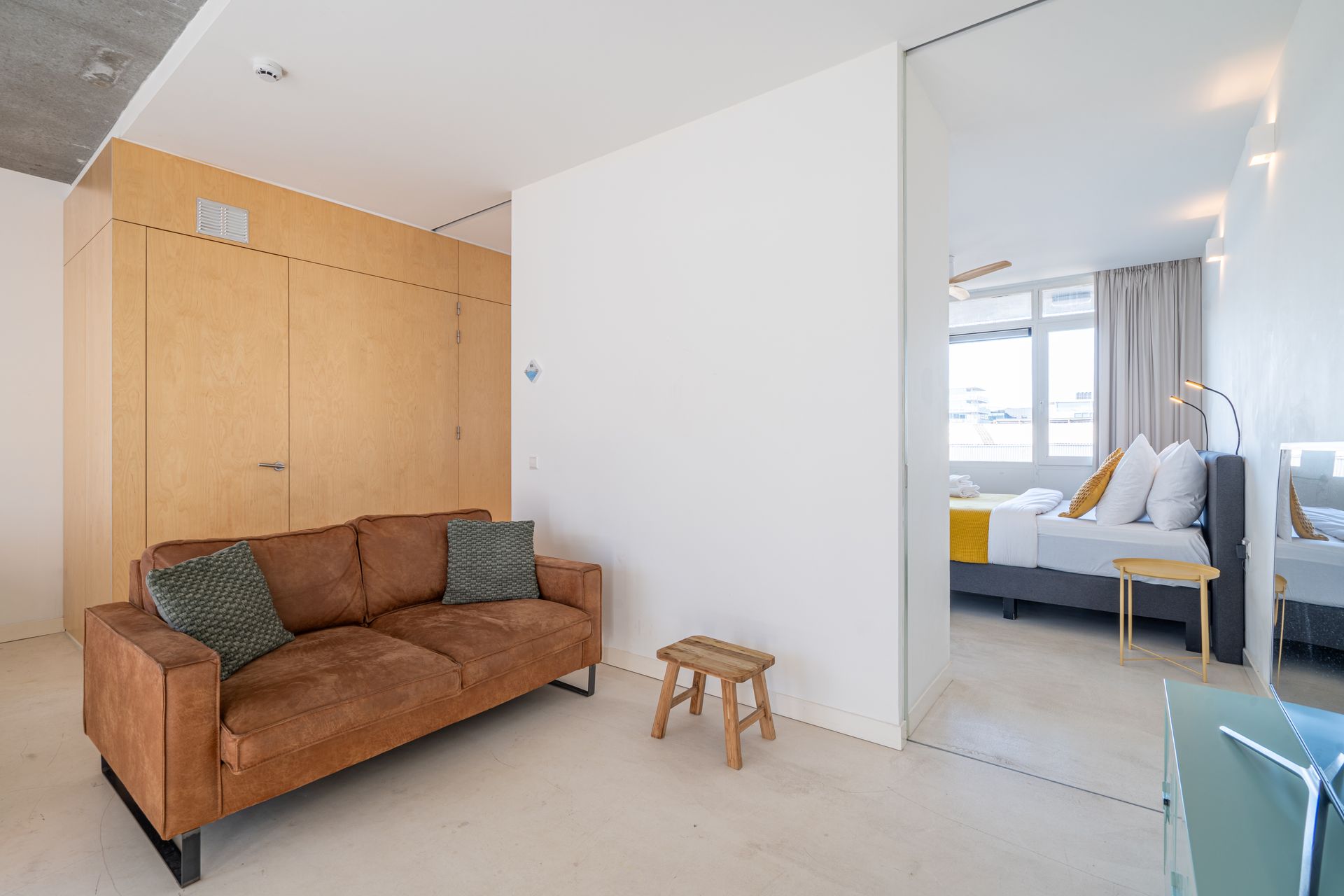 Een minimalistische woonkamer met een beige leren bank, een klein houten krukje en uitzicht op een slaapkamer met een bed.