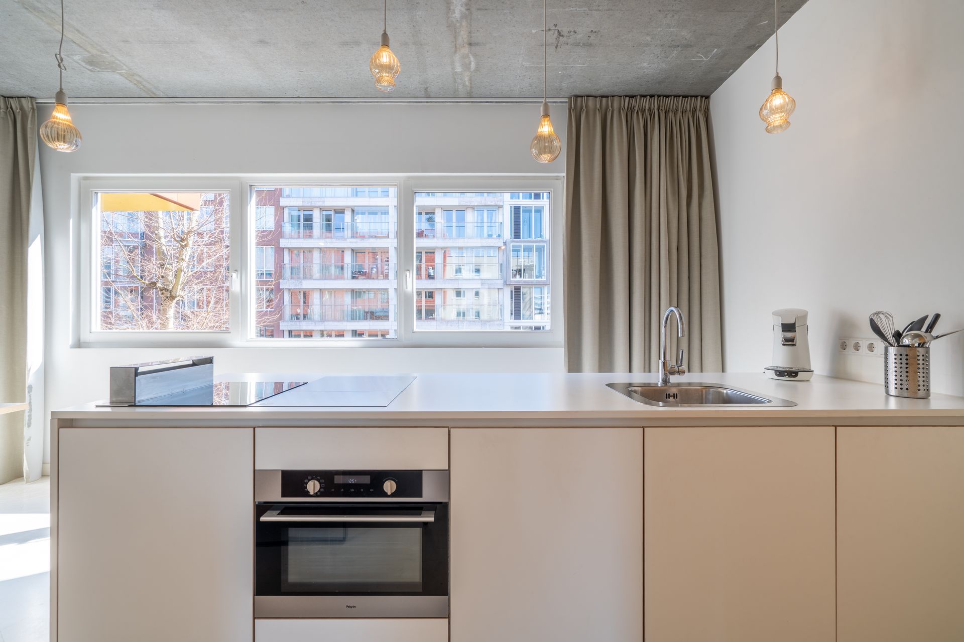 Modern wit keukeneiland met oven en spoelbak voor een raam, tegen een betonnen plafond en witte muren.