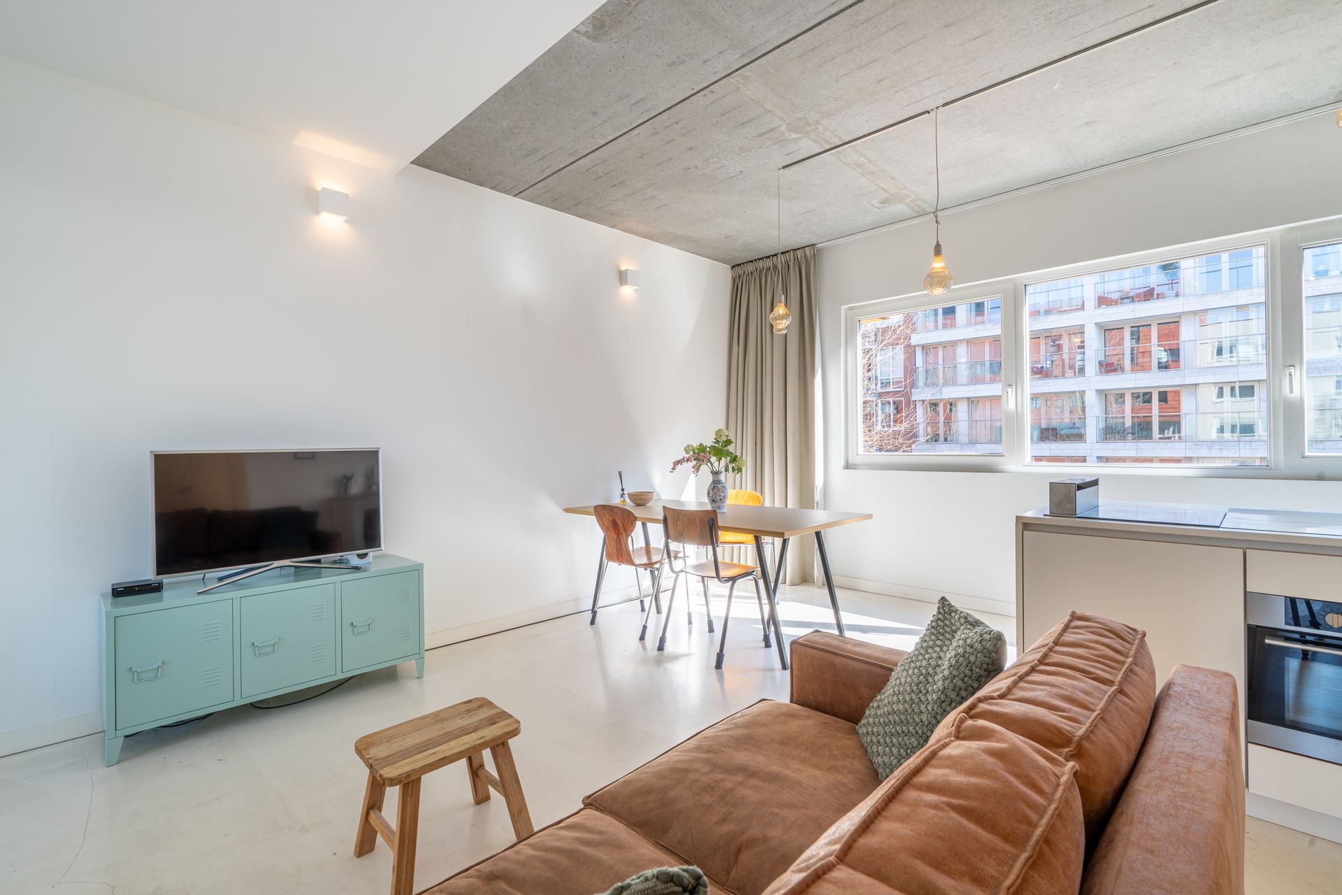 Een moderne woonkamer met een bruine bank, een blauw tv-meubel, een eethoek en een plafond van onbewerkt beton.
