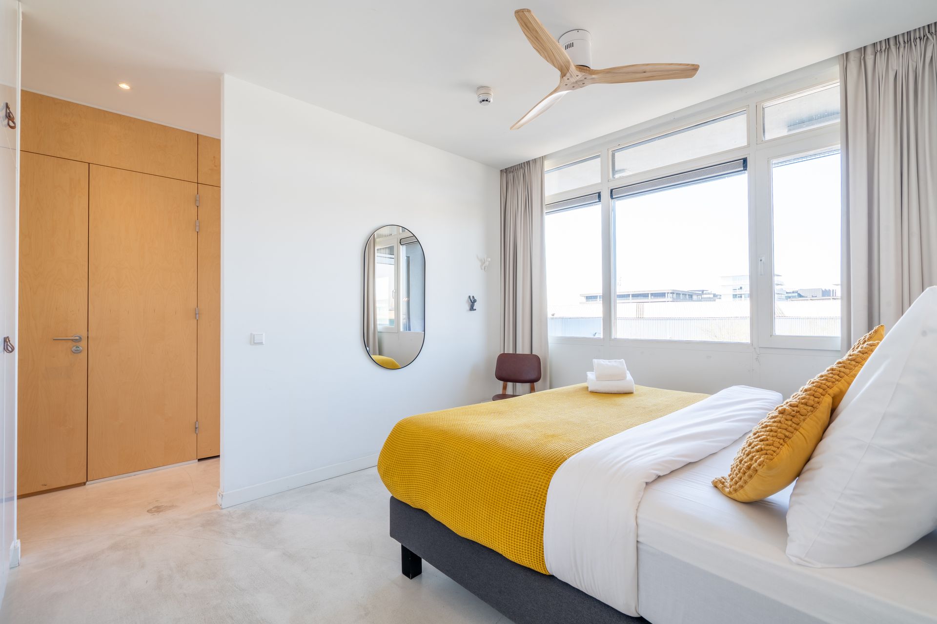 Een moderne slaapkamer met een bed met wit en geel beddengoed, een ronde spiegel, houten kastdeuren en een plafondventilator.