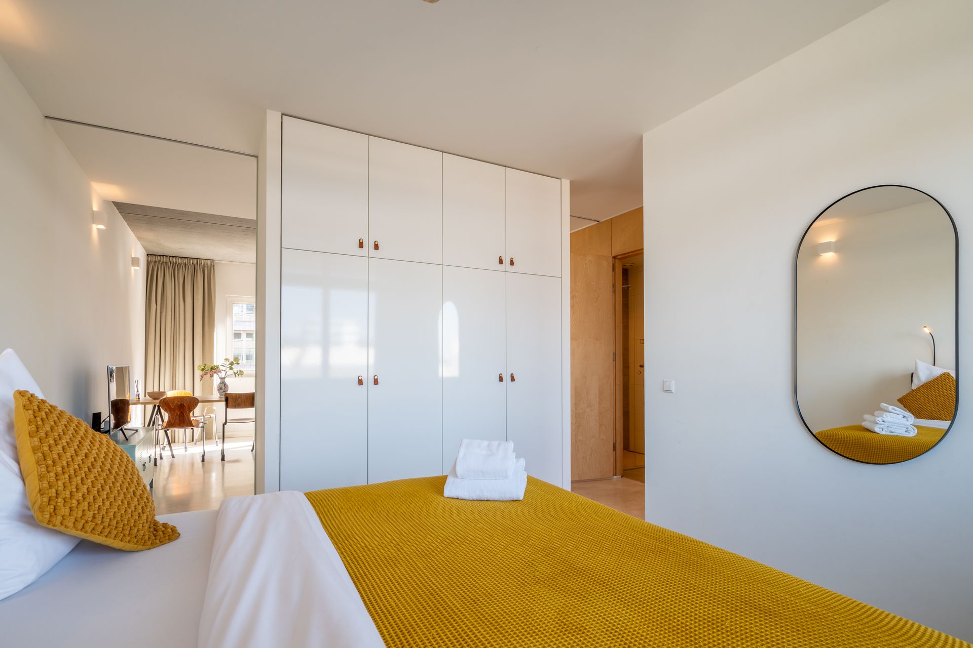 Een slaapkamer met een geel gewatteerd dekbed, een witte kledingkast en een ovale spiegel aan een effen muur.