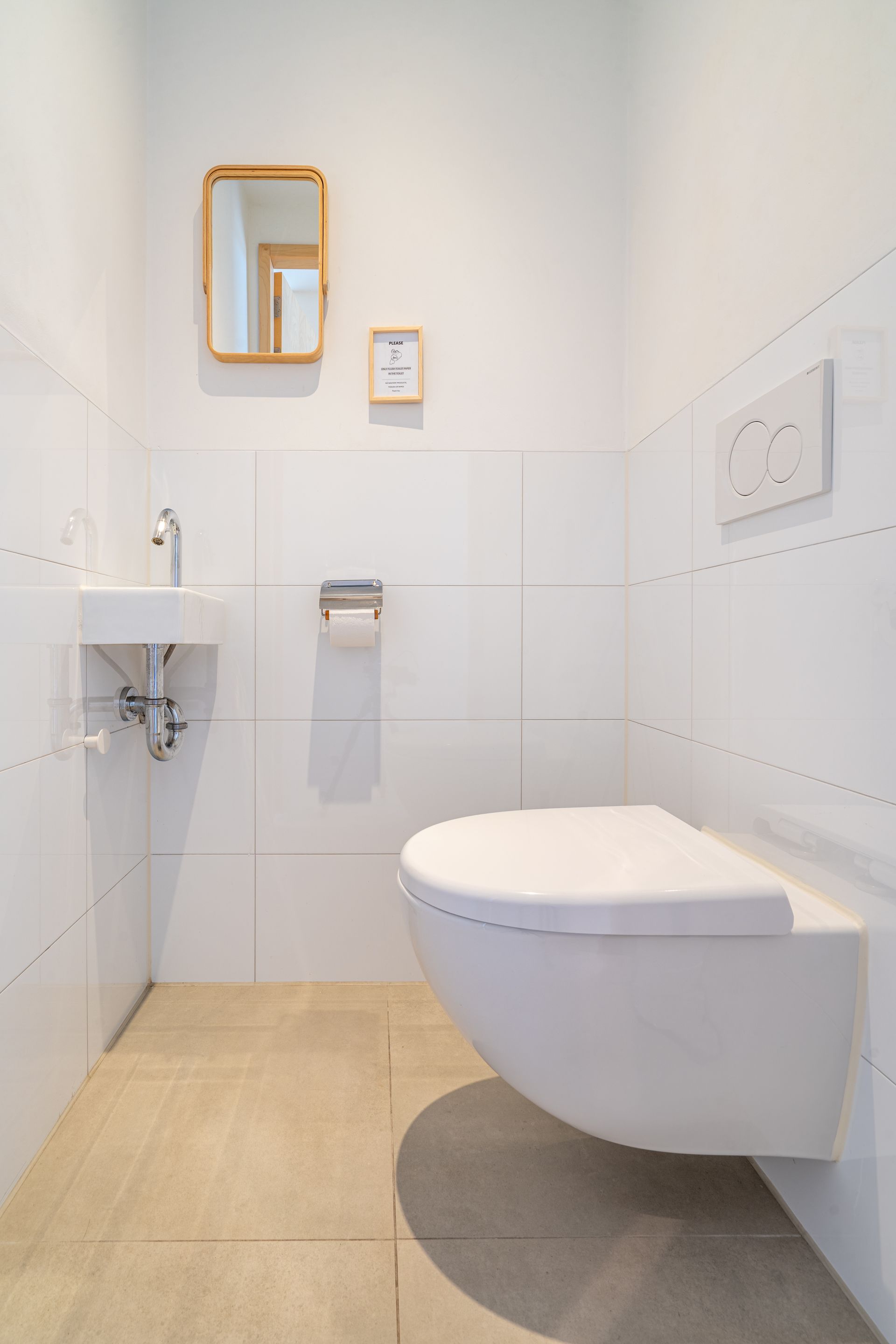 Een minimalistische witte badkamer met een wandtoilet, een kleine wastafel, een spiegel met gouden lijst en beige tegelvloer.