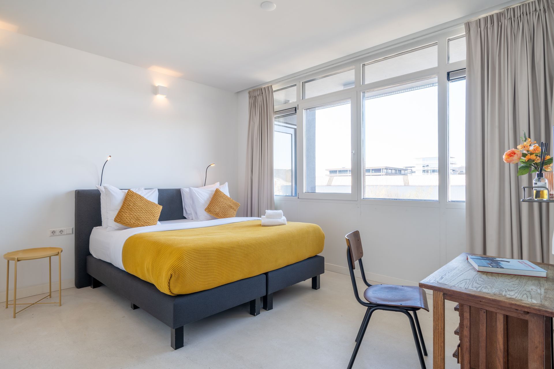 Een moderne hotelkamer met een bed met donker frame, felgeel beddengoed, een houten bureau met stoel en grote ramen.