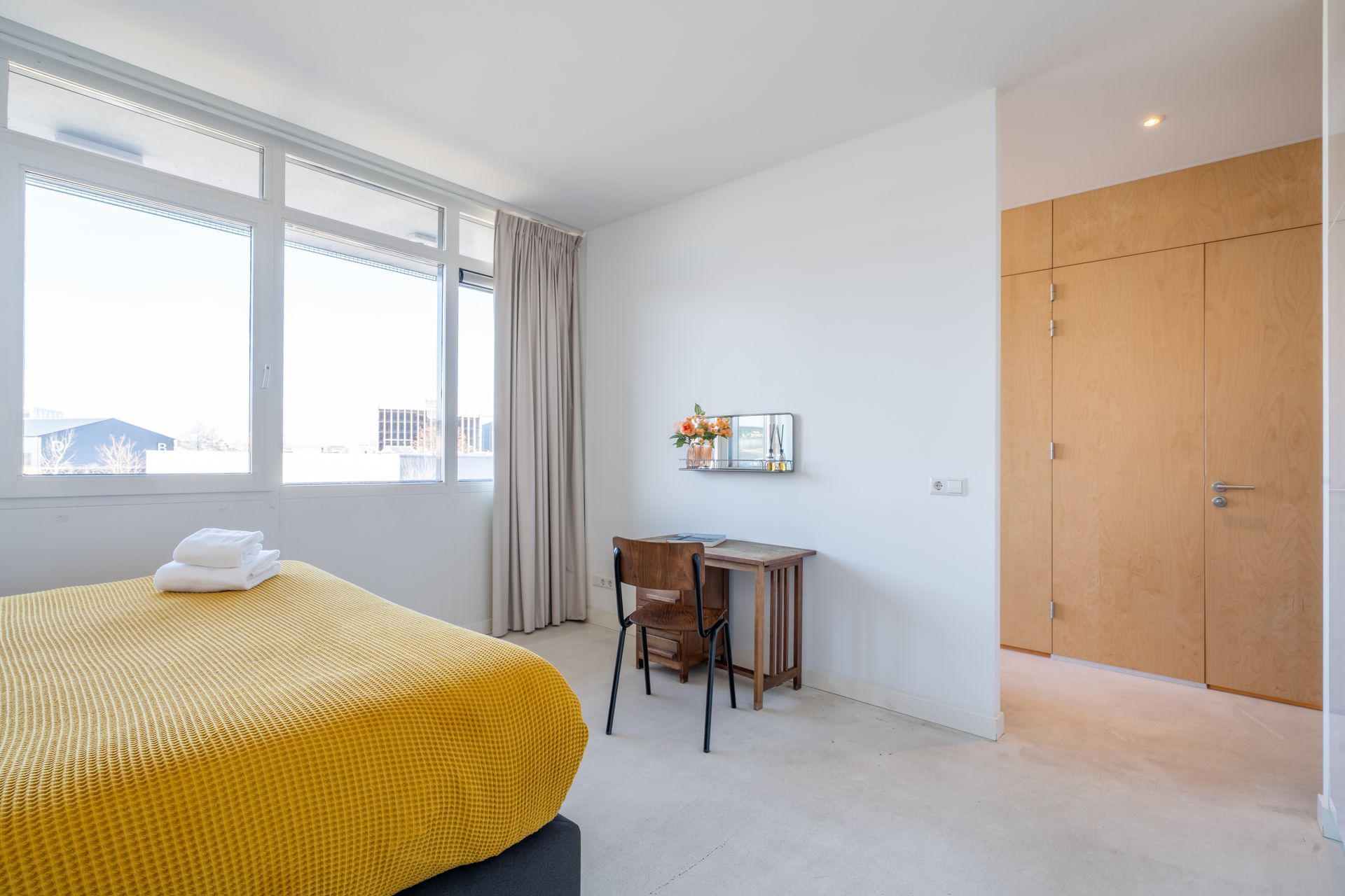 Een slaapkamer met een geel bed, een houten bureau met een stoel en grote ramen met kamerhoge gordijnen bij een houten deur.