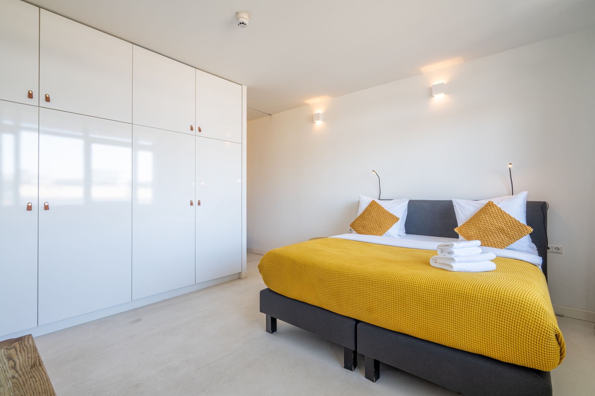 Een hotelkamer met een bed voorzien van een felgele gebreide plaid en bijpassende kussens, naast grote witte kledingkasten.