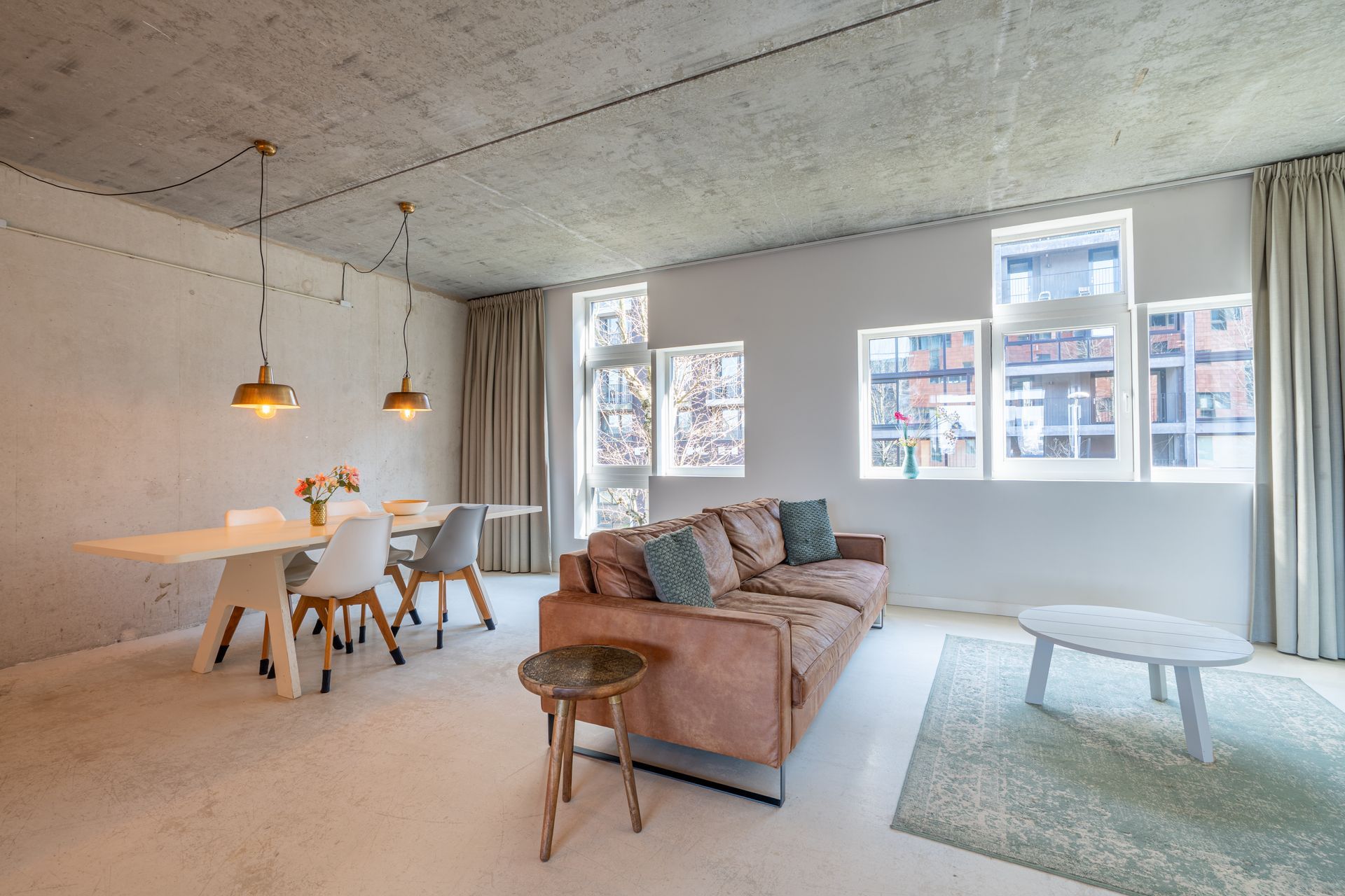 Een minimalistische woonkamer met een bruine bank, een eettafel met stoelen, een betonnen plafond en grote ramen.