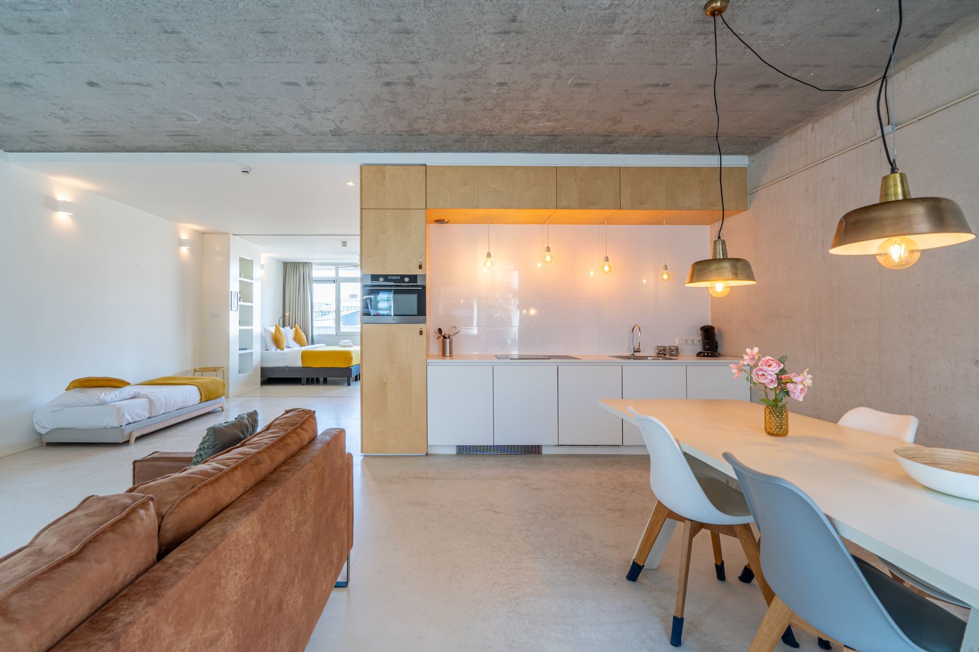 Een moderne, open woonruimte met een keuken, eethoek, bruine bank en een slaapkamer op de achtergrond.