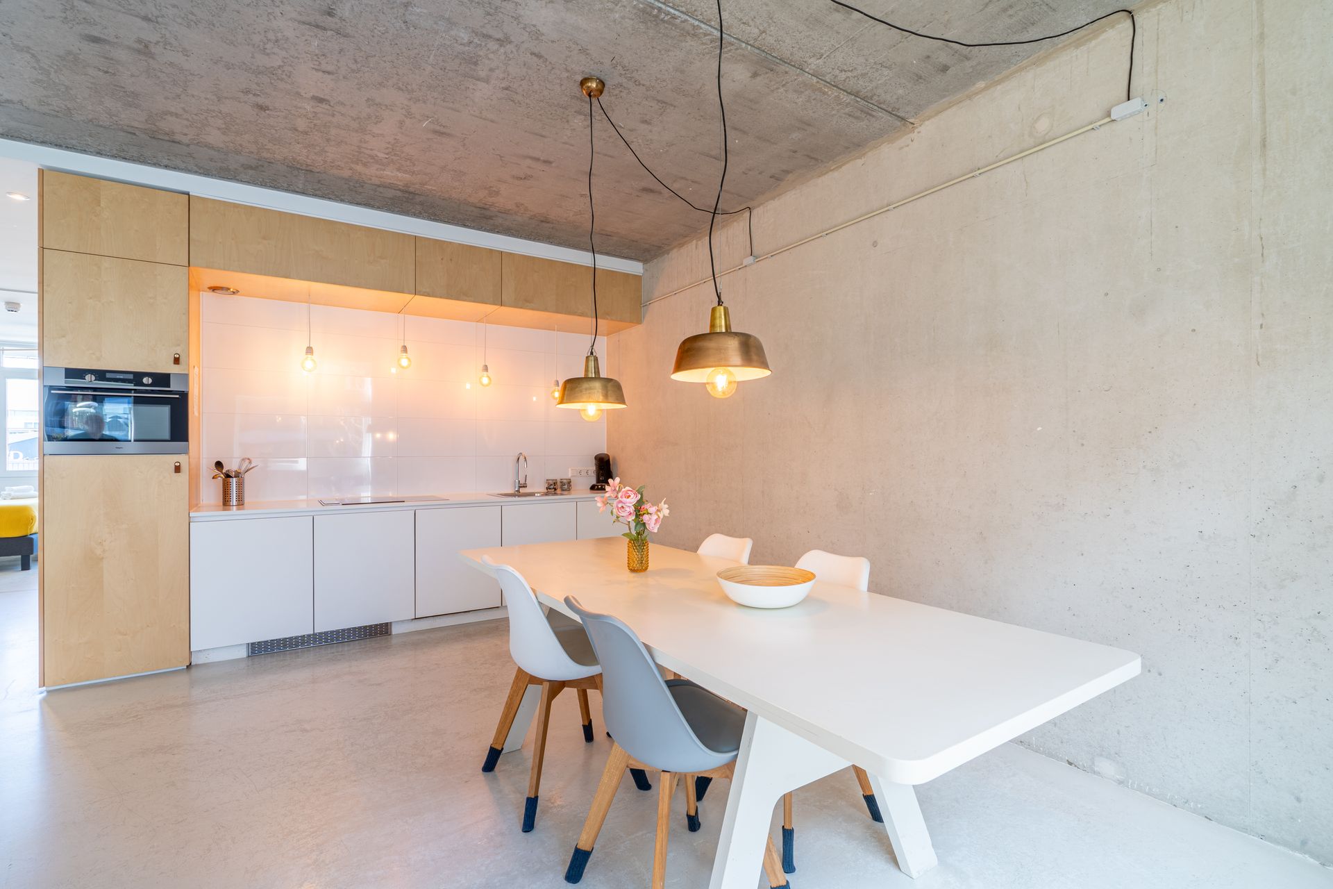Een minimalistische eethoek met een witte tafel en stoelen voor een keuken met lichte houten kasten en een betonnen muur.