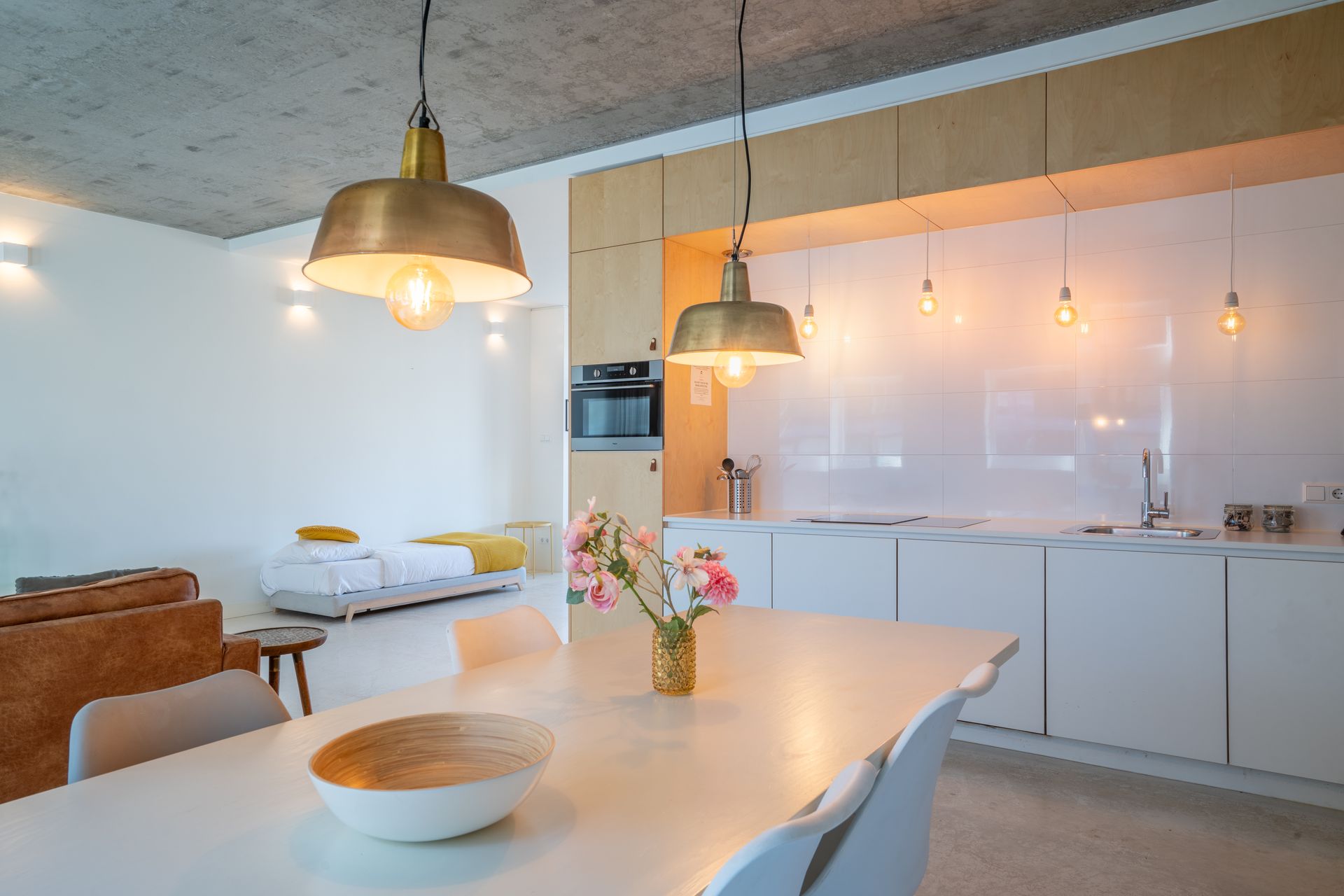 Een modern, open appartement met een witte eettafel, lichte houten keukenkasten, een betonnen plafond en gouden hanglampen.