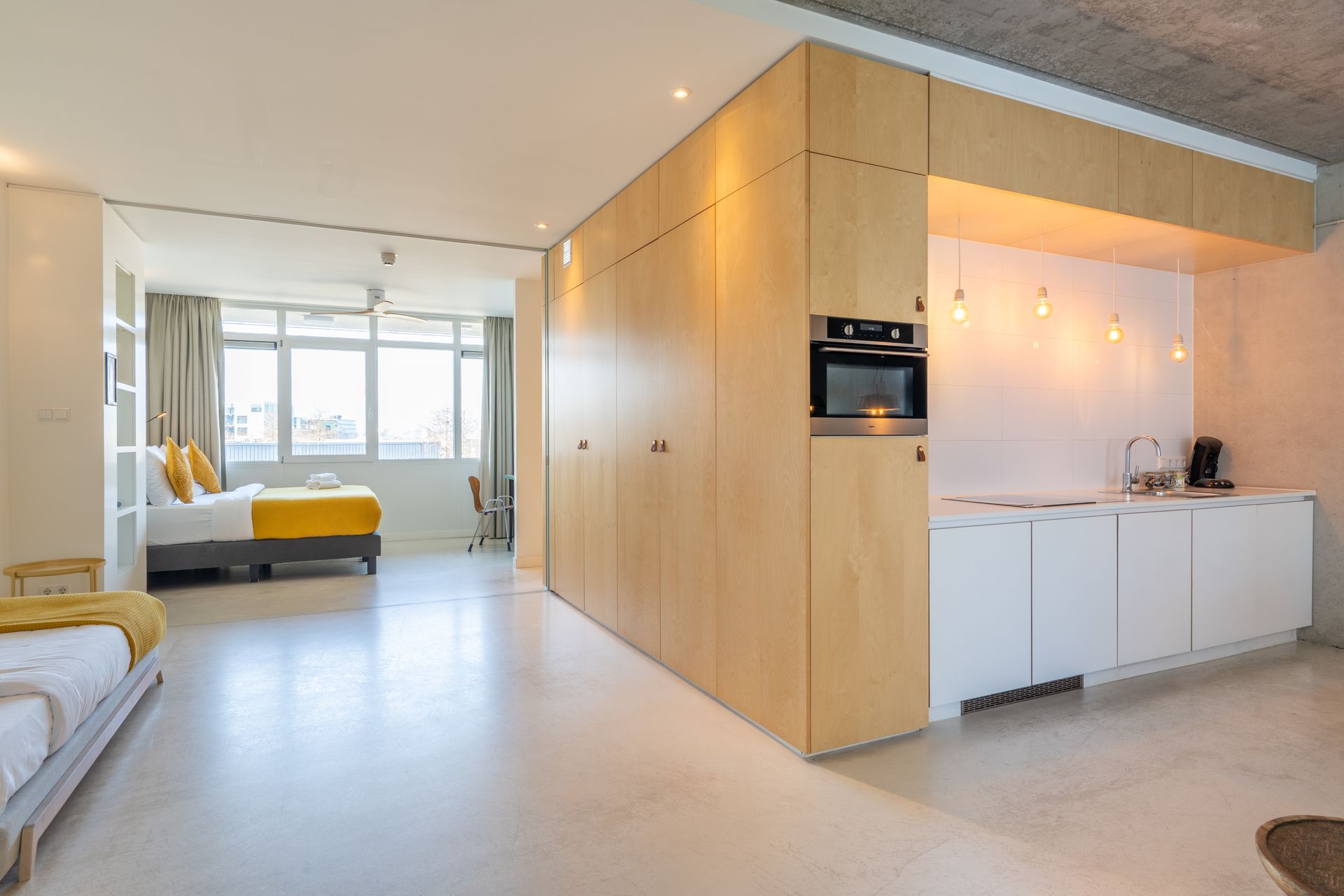 Een modern, open studio-appartement met een bed, een minimalistische keuken met lichte houten kasten en witte vloeren.