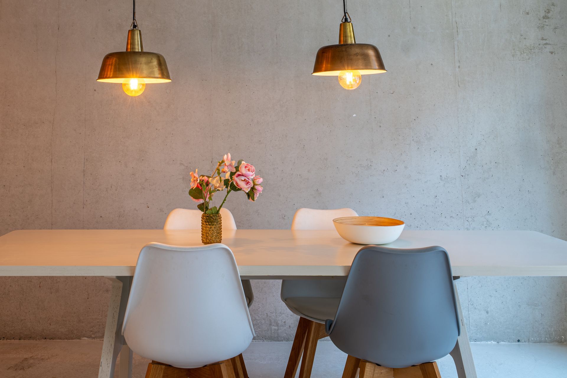 Een witte eettafel met twee stoelen, een vaas met roze bloemen en een schaal tegen een effen, gestructureerde betonnen muur.