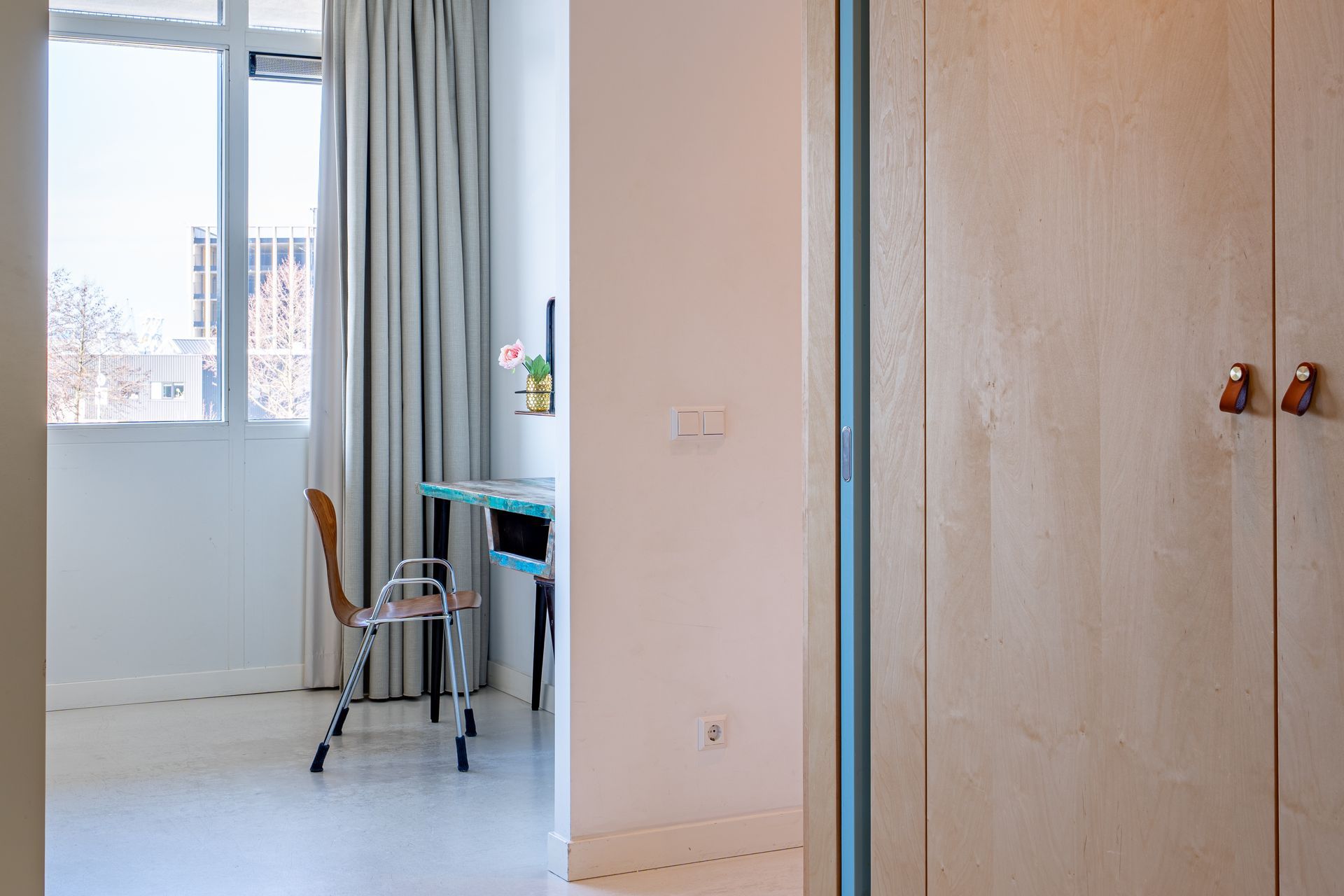 Een blik door een deuropening naar een lichte kamer met een bureau, een houten stoel en gordijnen voor een raam.