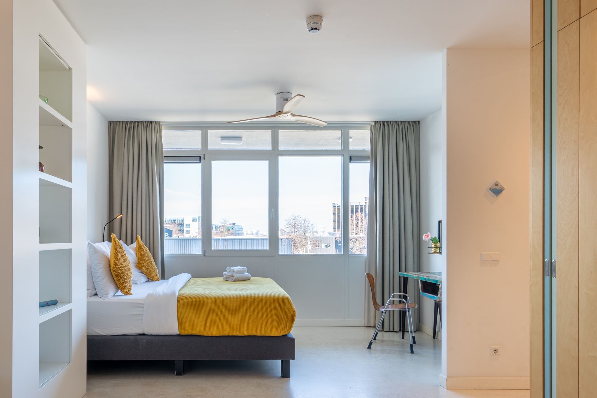 Een hotelkamer met een tweepersoonsbed, een gele plaid, lichtgrijze gordijnen, een klein bureau