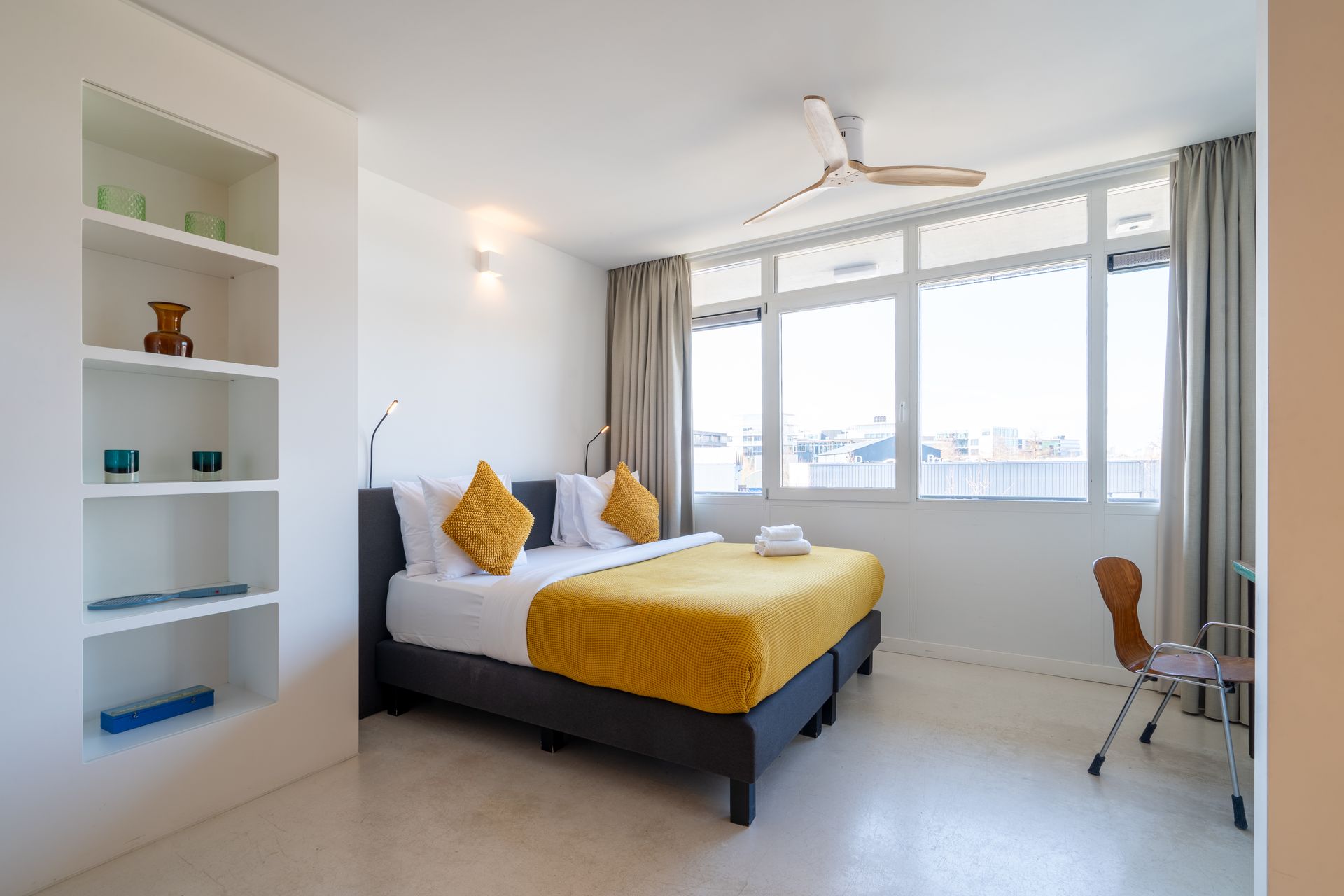 Een hotelkamer met een tweepersoonsbed, een gele plaid, wit beddengoed, open planken en een bureaustoel.