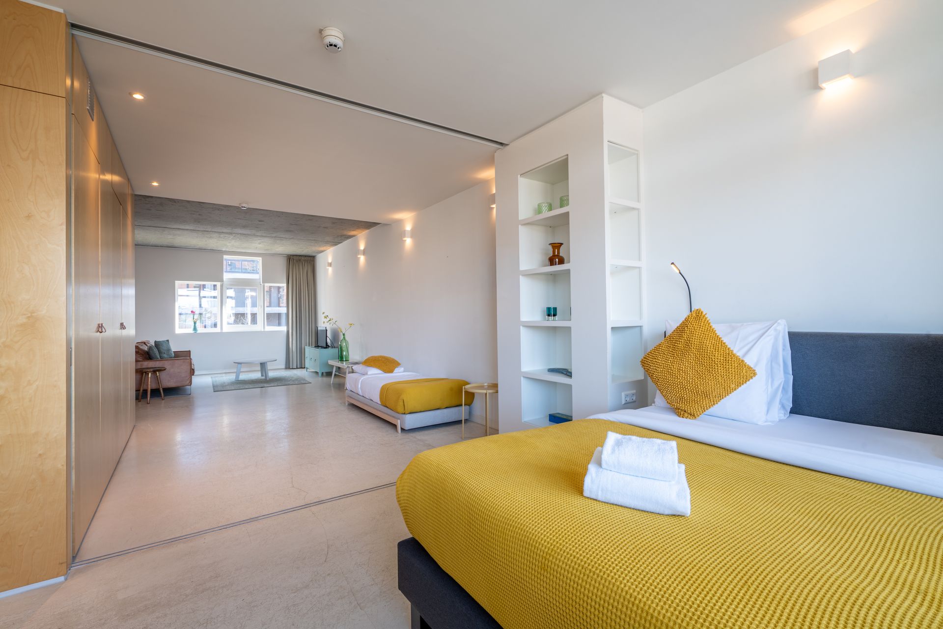 Een hotelkamer met twee bedden, geel beddengoed met een patroon, witte muren en een hoge boekenkast.