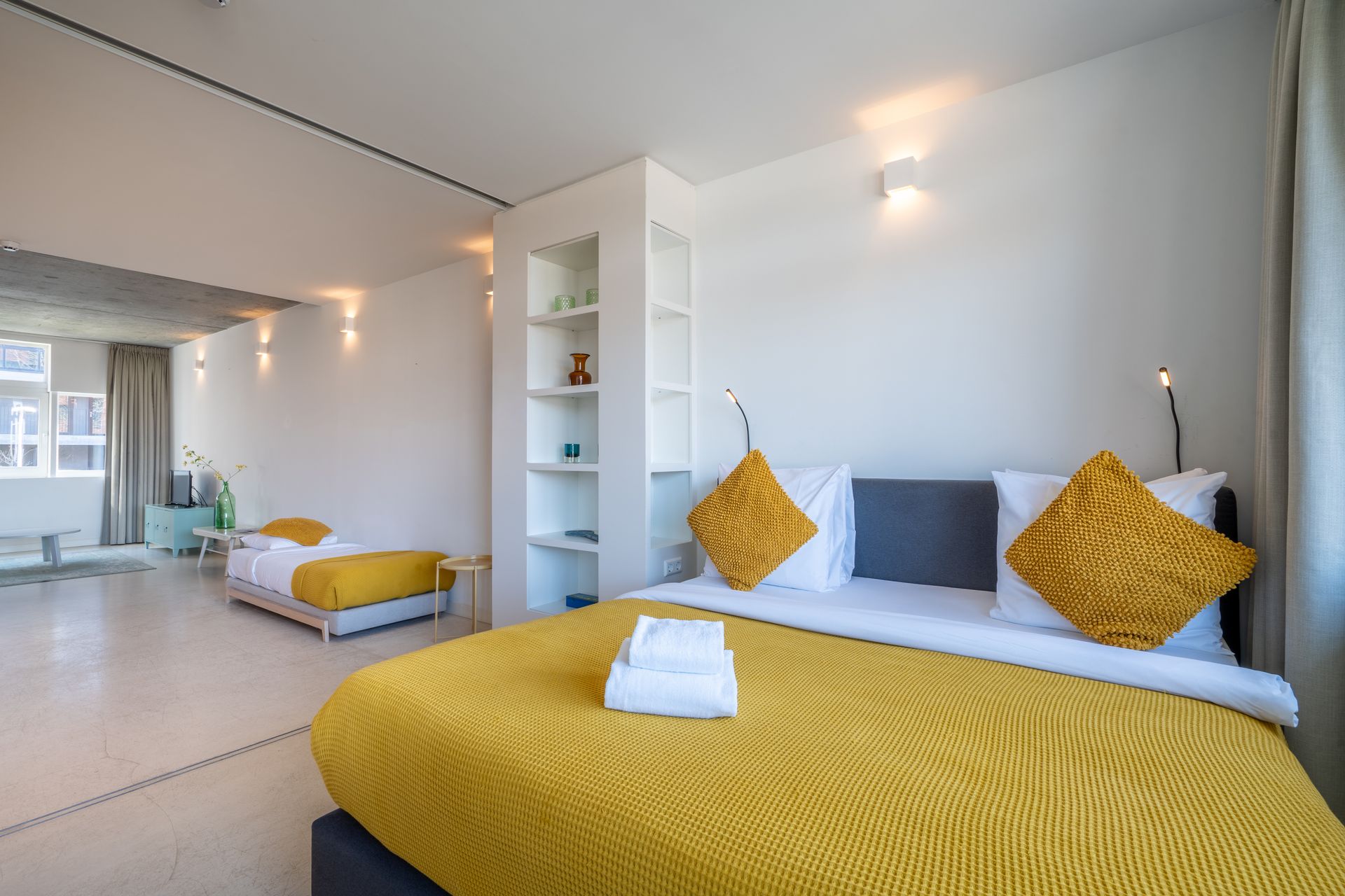 Een lichte hotelkamer met een groot bed op de voorgrond en een tweede bed op de achtergrond, met gele accenten.