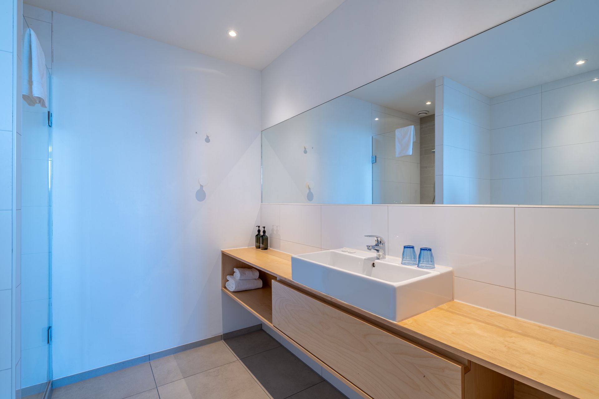 Moderne, minimalistische badkamer met een witte wastafel op een licht houten badmeubel, een grote spiegel en betegelde muren.