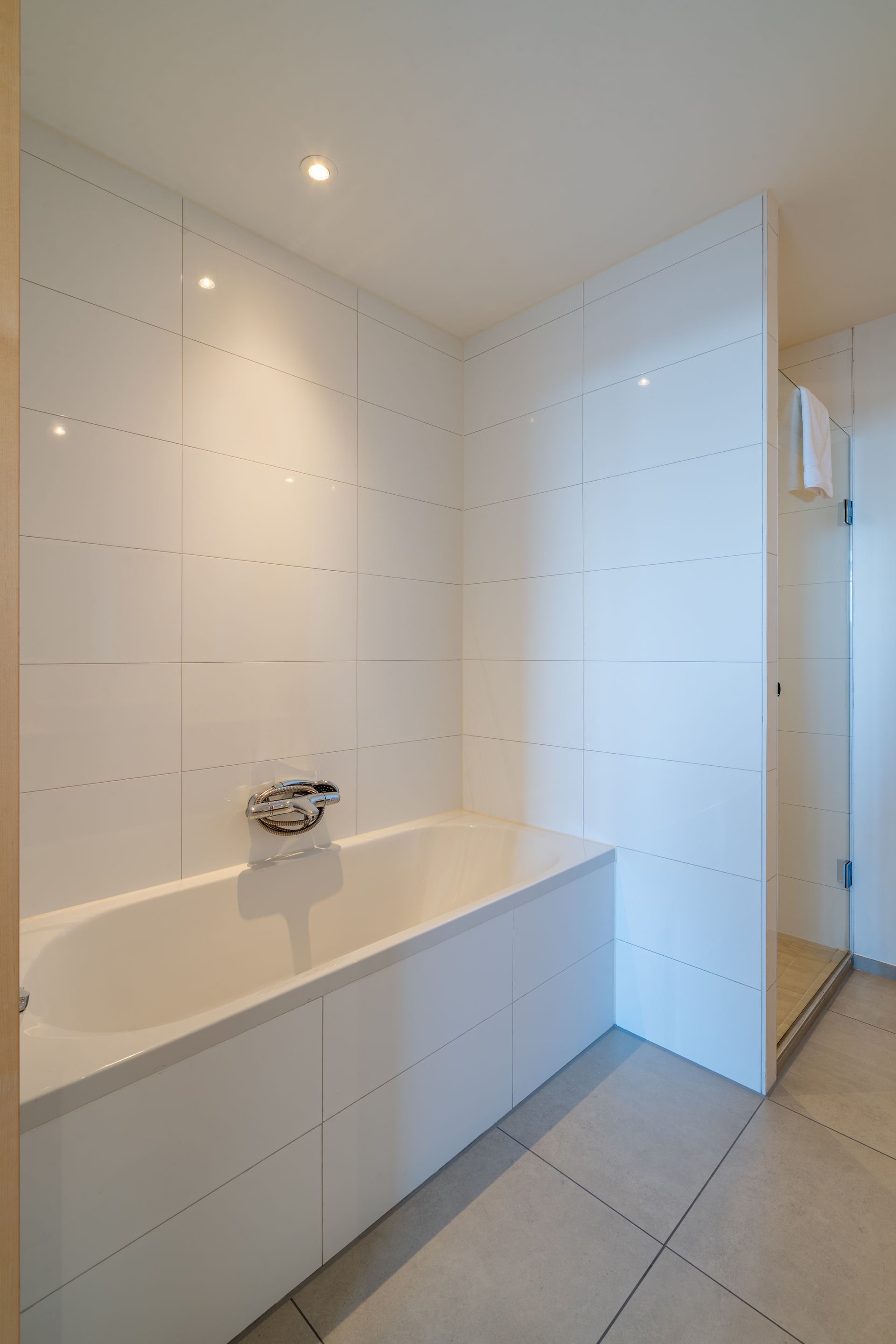 Een moderne witte badkamer met een ingebouwd bad tegen een betegelde muur en een glazen douchecabine ernaast.