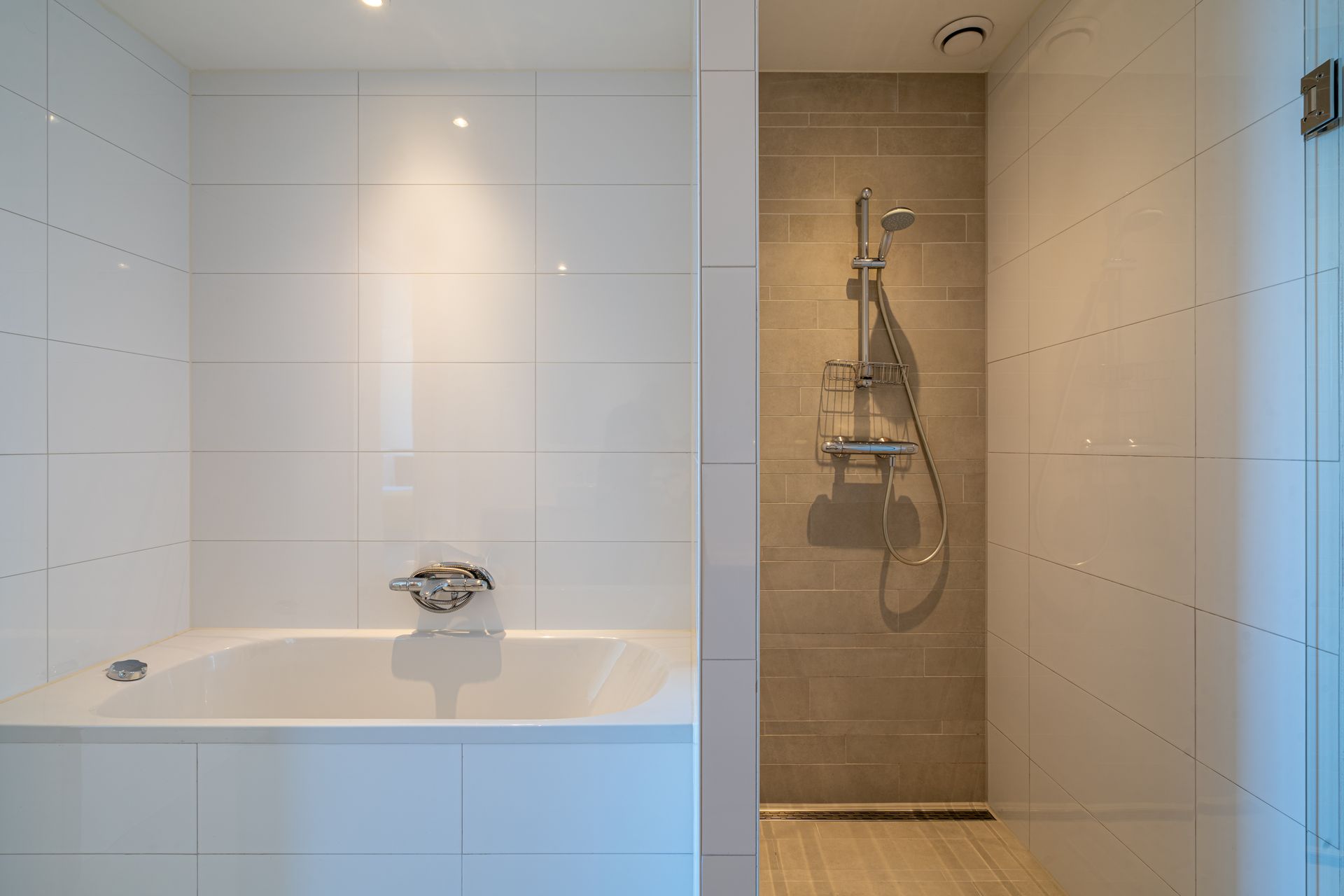 Een moderne badkamer met een wit bad naast een aparte, glazen douchecabine met lichtbeige, gestructureerde tegels.