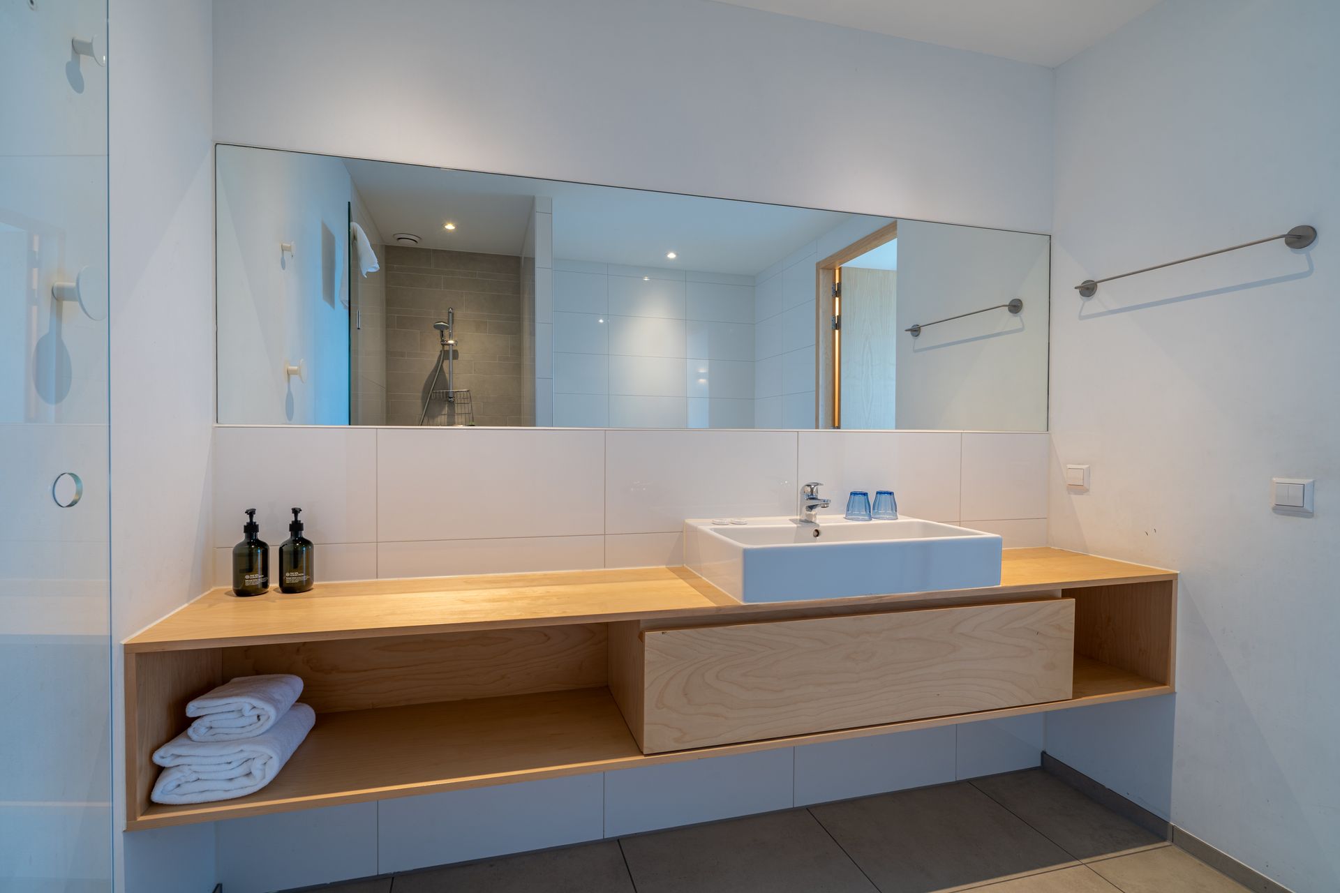 Moderne badkamerkast met een witte wasbak, een grote spiegel en open houten planken met opgevouwen witte handdoeken.