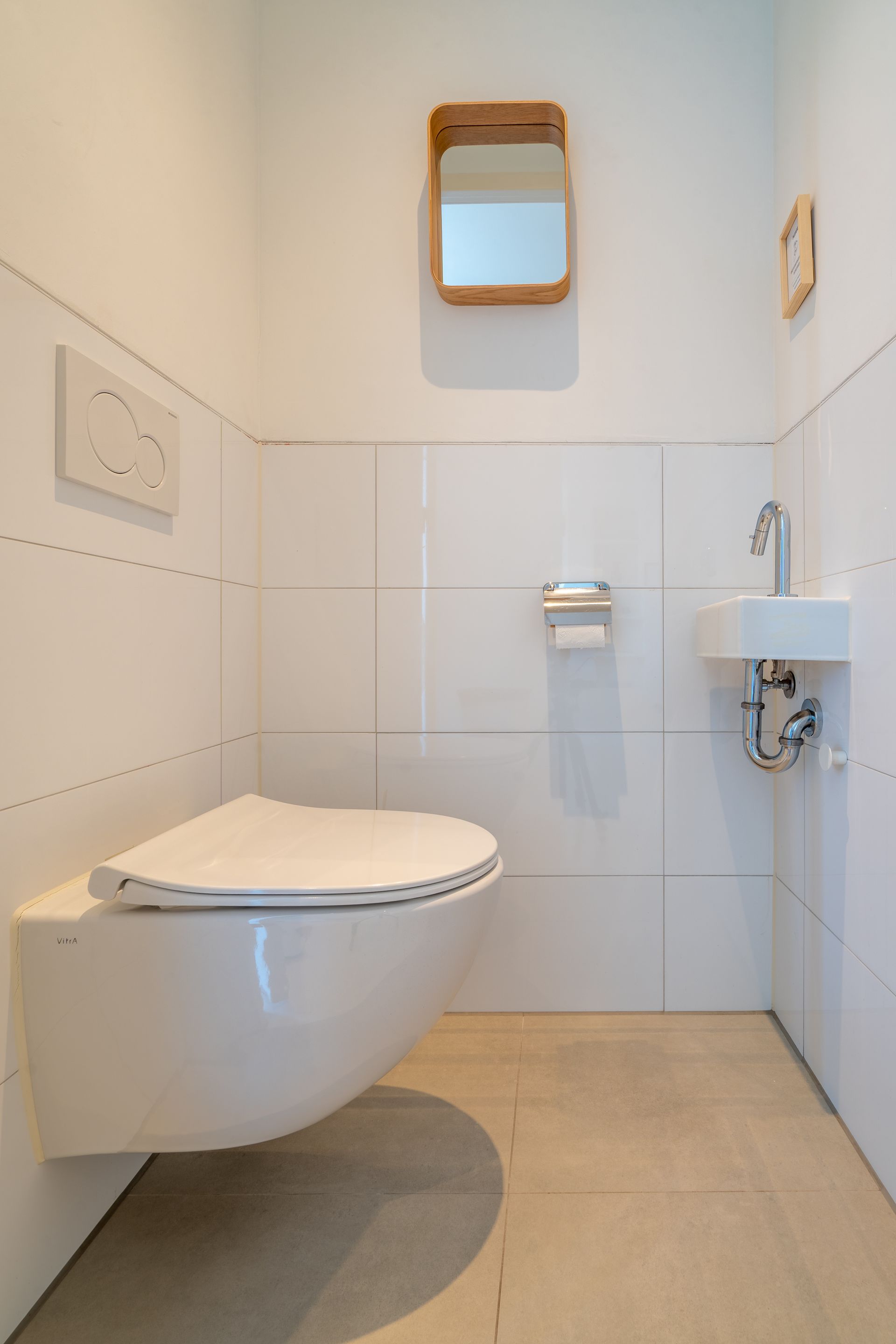 Een modern wit wandtoilet, een kleine wastafel met een chromen kraan en een kleine houten spiegel in een betegelde badkamer.