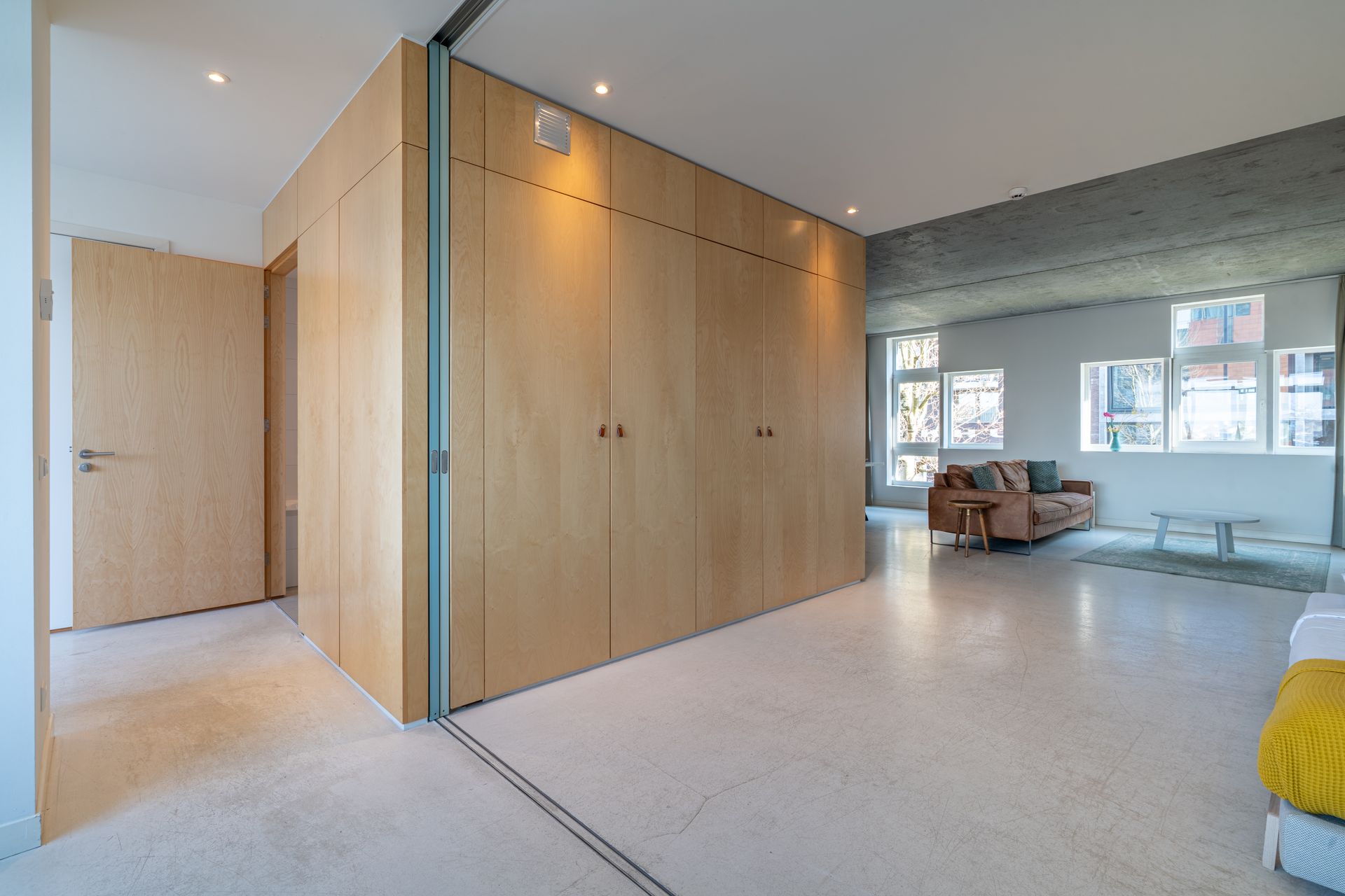 Modern interieur met lichte houten kasten, witte vloer en een zithoek met een bank op de achtergrond.