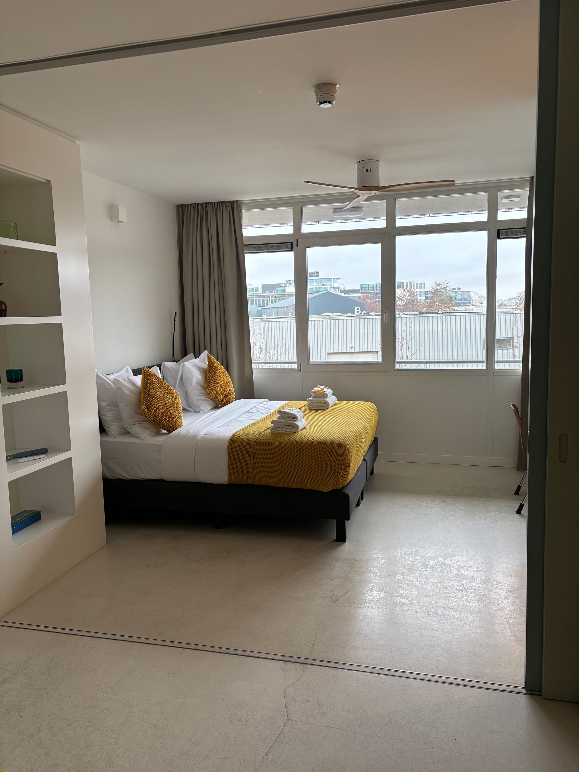 Slaapkamer met bed, kussens, raam en ingebouwde planken. Witte muren, grijze gordijnen en een gele sprei.