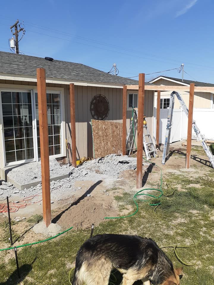 Construction of a patio cover: wooden posts installed along a house, gravel ground, German Shepherd in foreground.