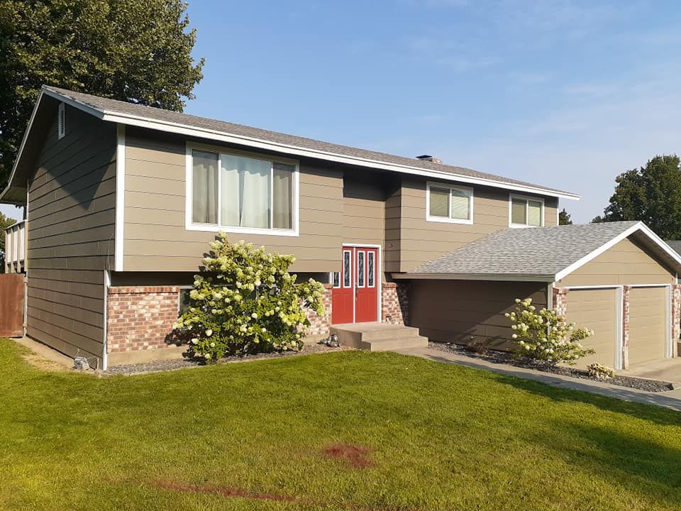 Two-story house with green siding, red door, and garage. Lush green lawn and bushes in front.