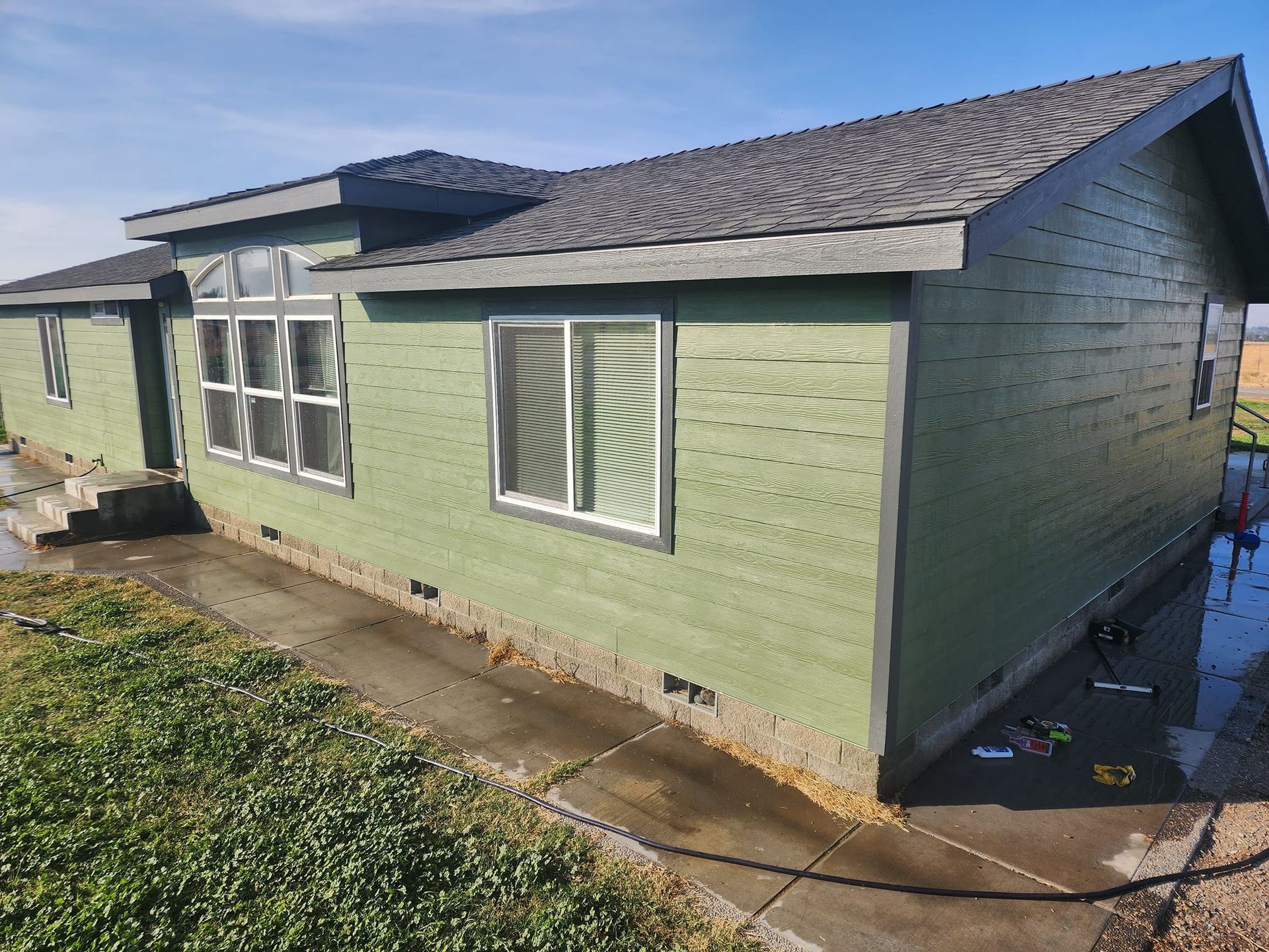Green-sided house with a dark gray roof and white-trimmed windows on a sunny day.