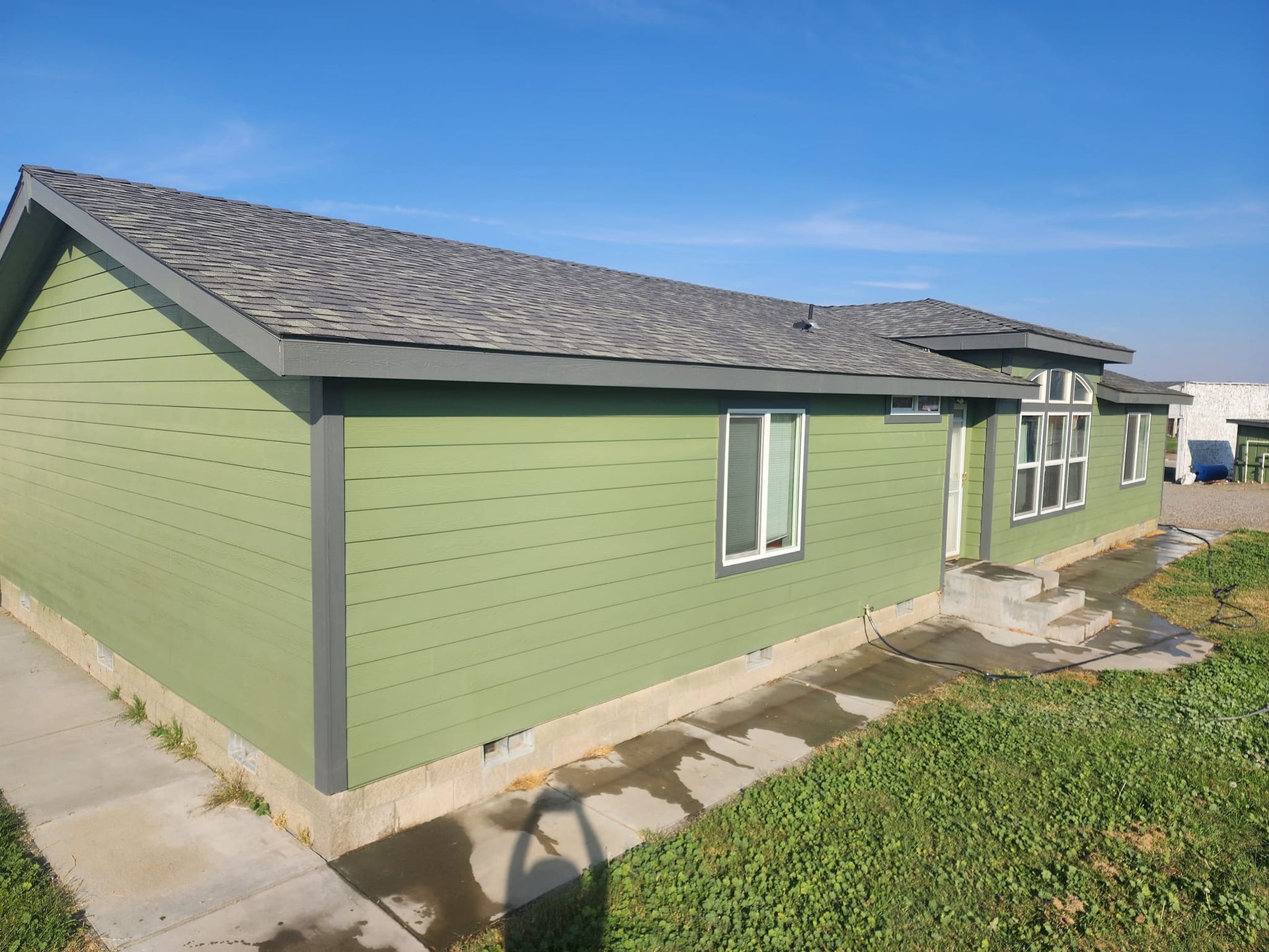 Green house with gray roof, white window trim, and a concrete path on a sunny day.