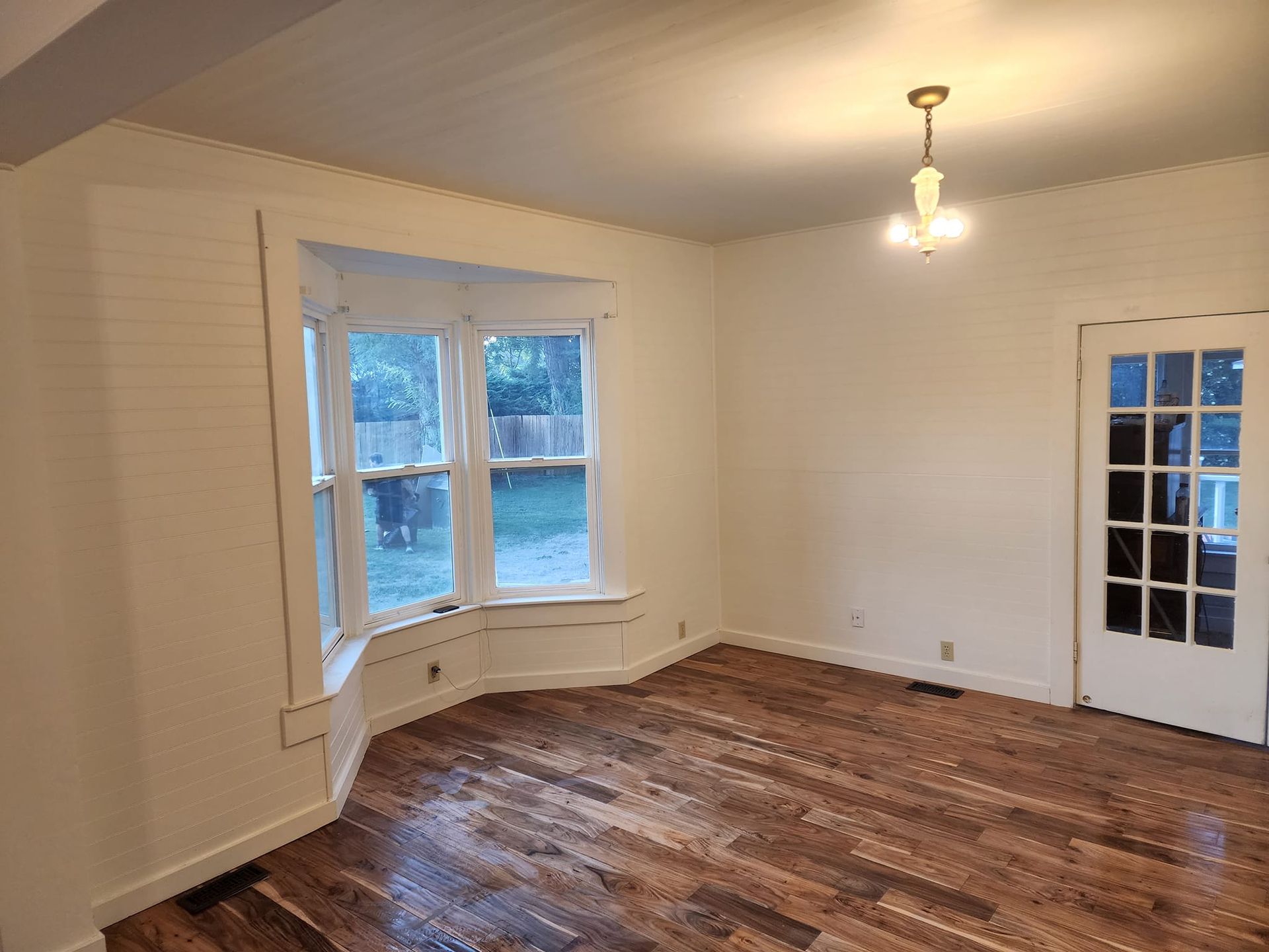 Empty room with hardwood floors, a bay window, and a door; walls are white.