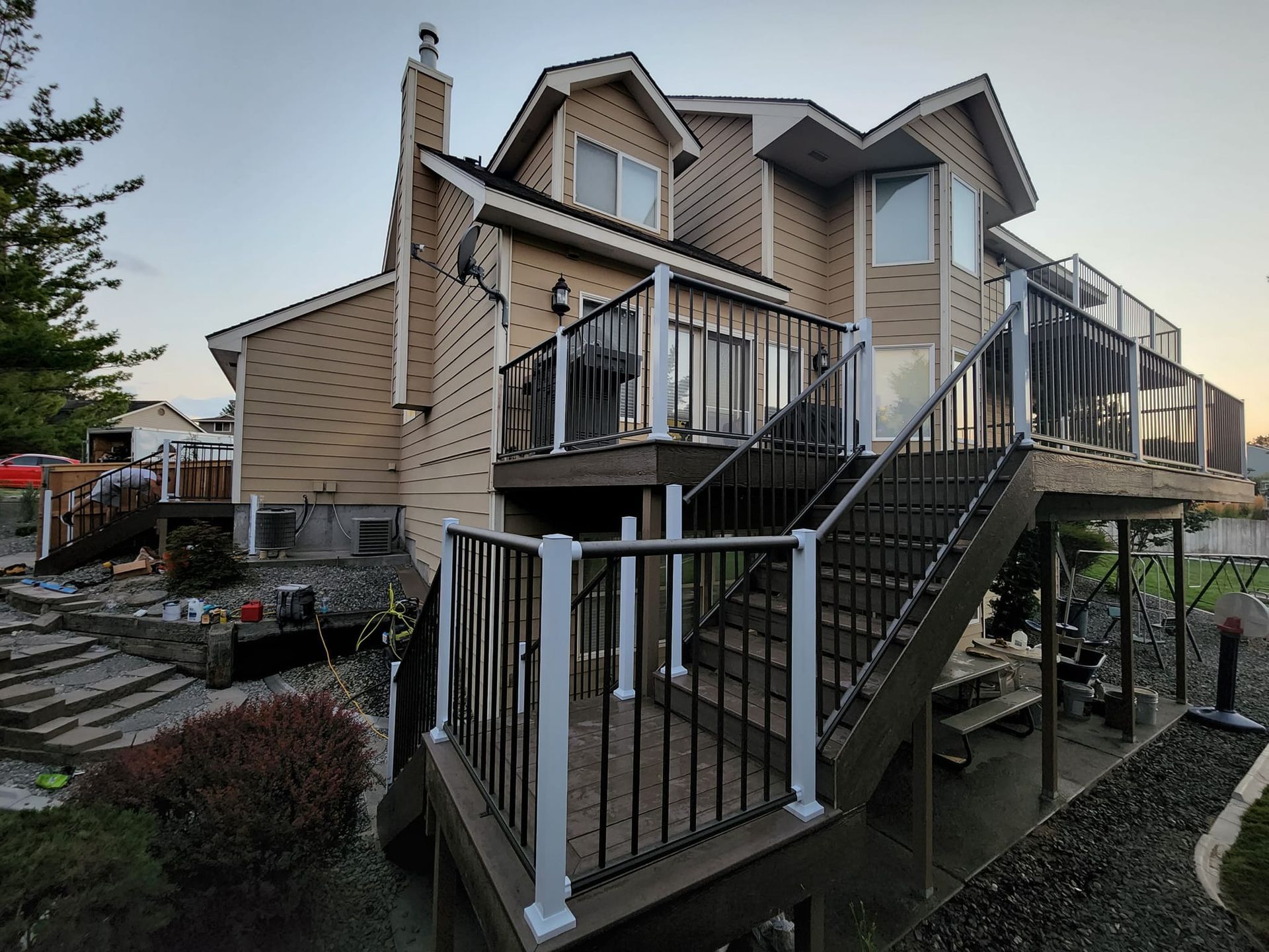 Two-story beige house with deck and black railing. Staircase leads down to yard with dark deck and bushes.