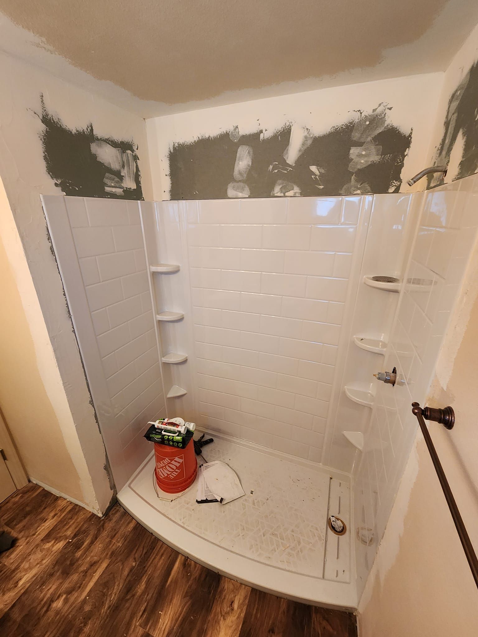 New white shower stall installation in a bathroom with wood-look flooring and unfinished walls.