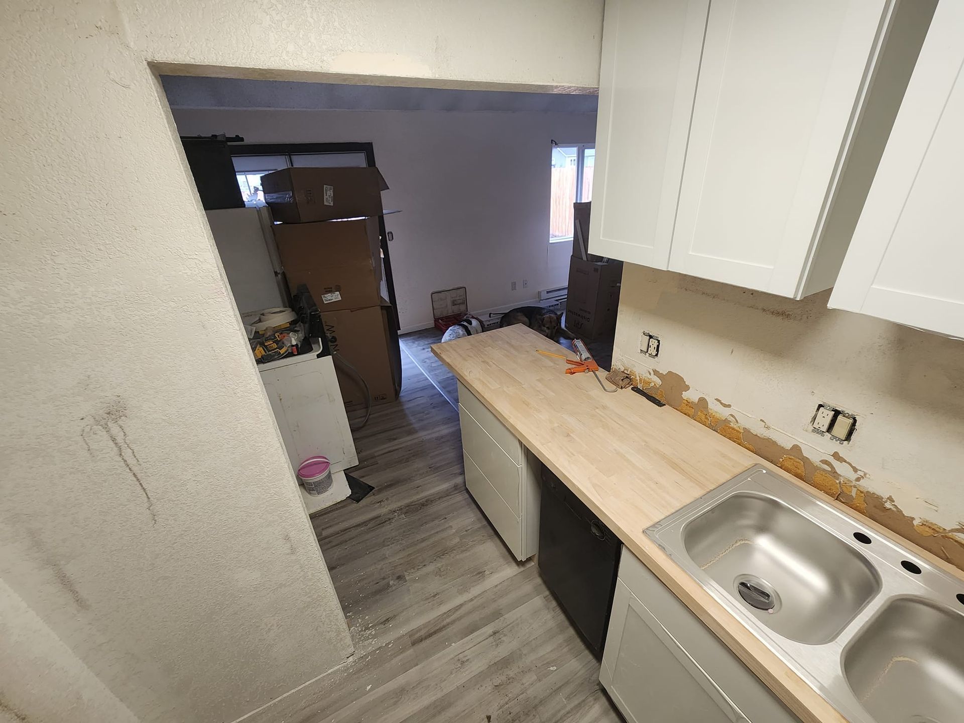 Kitchen undergoing renovation; white cabinets, new countertops, exposed wall. View into cluttered room.