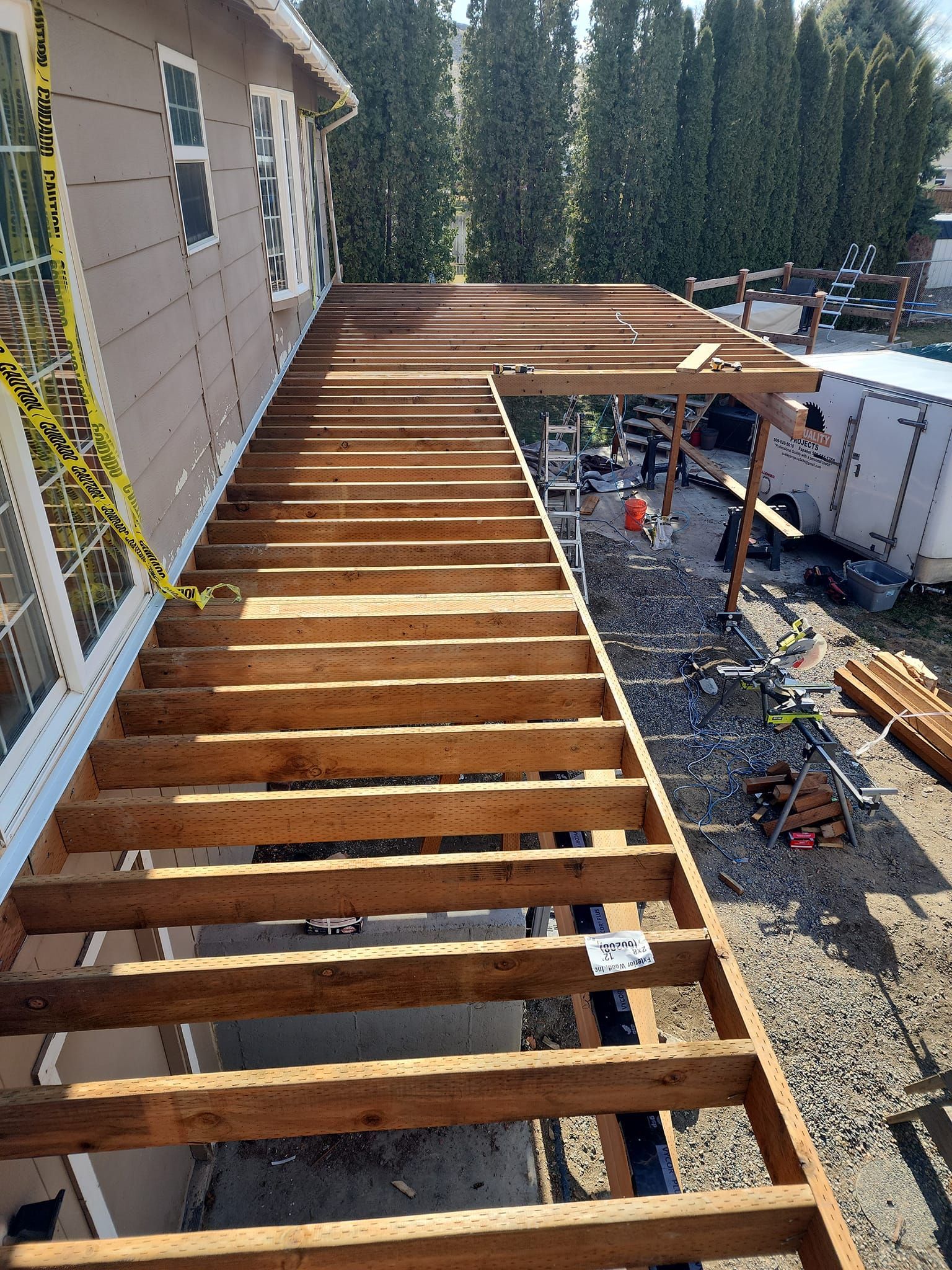 Wooden deck under construction next to a house with a small roofed section. Gravel ground.