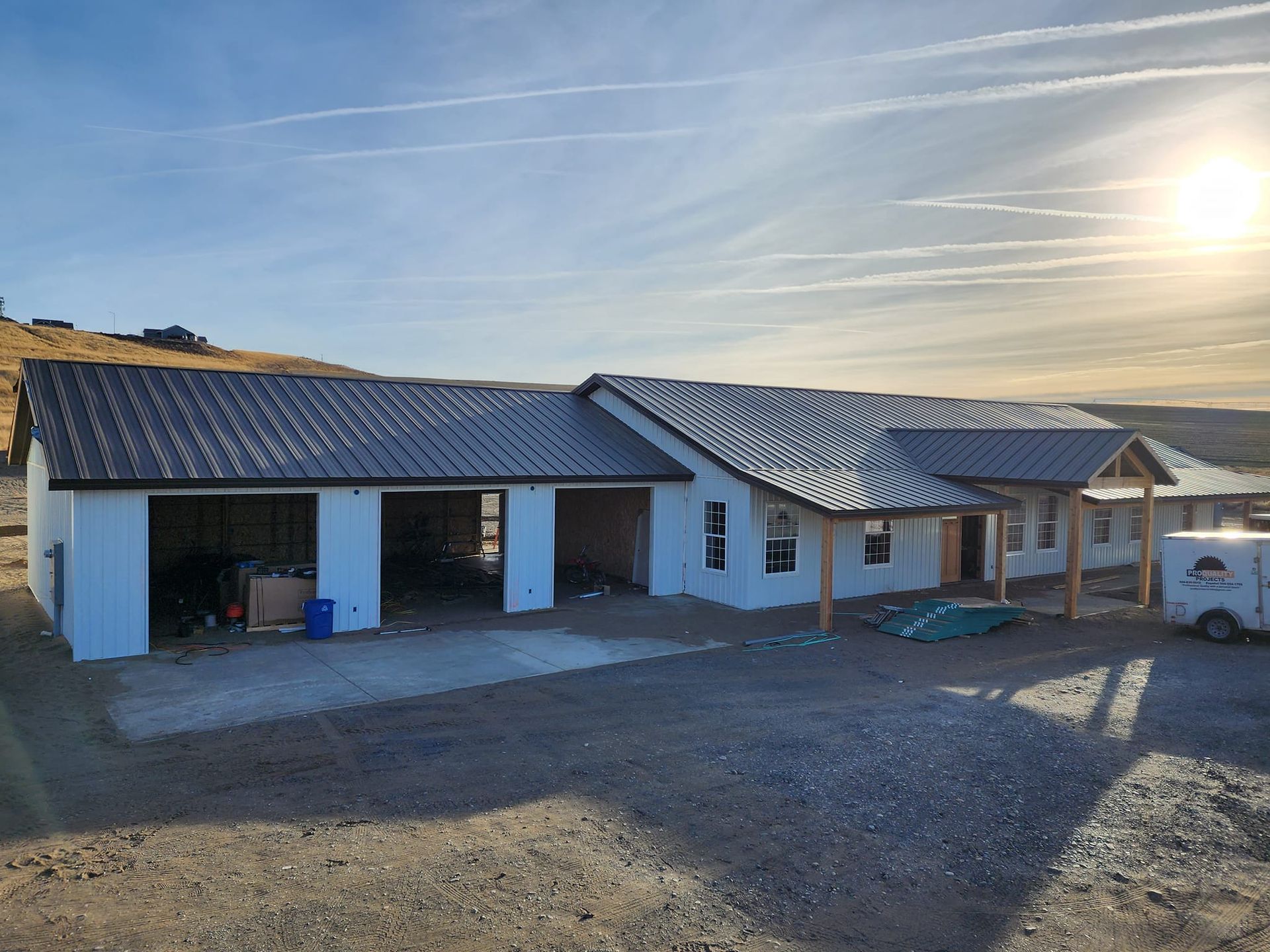 House under construction with garage, white walls, gray roof, on a gravel lot under a sunny sky.