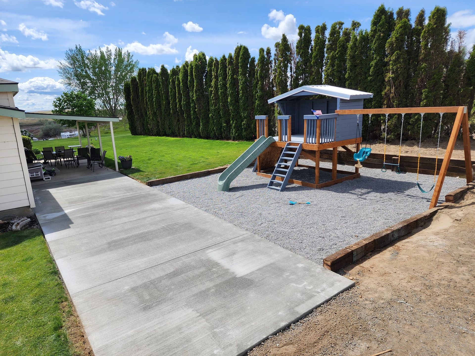 Backyard with concrete patio, gravel play area, wooden playhouse with slide and swing set.