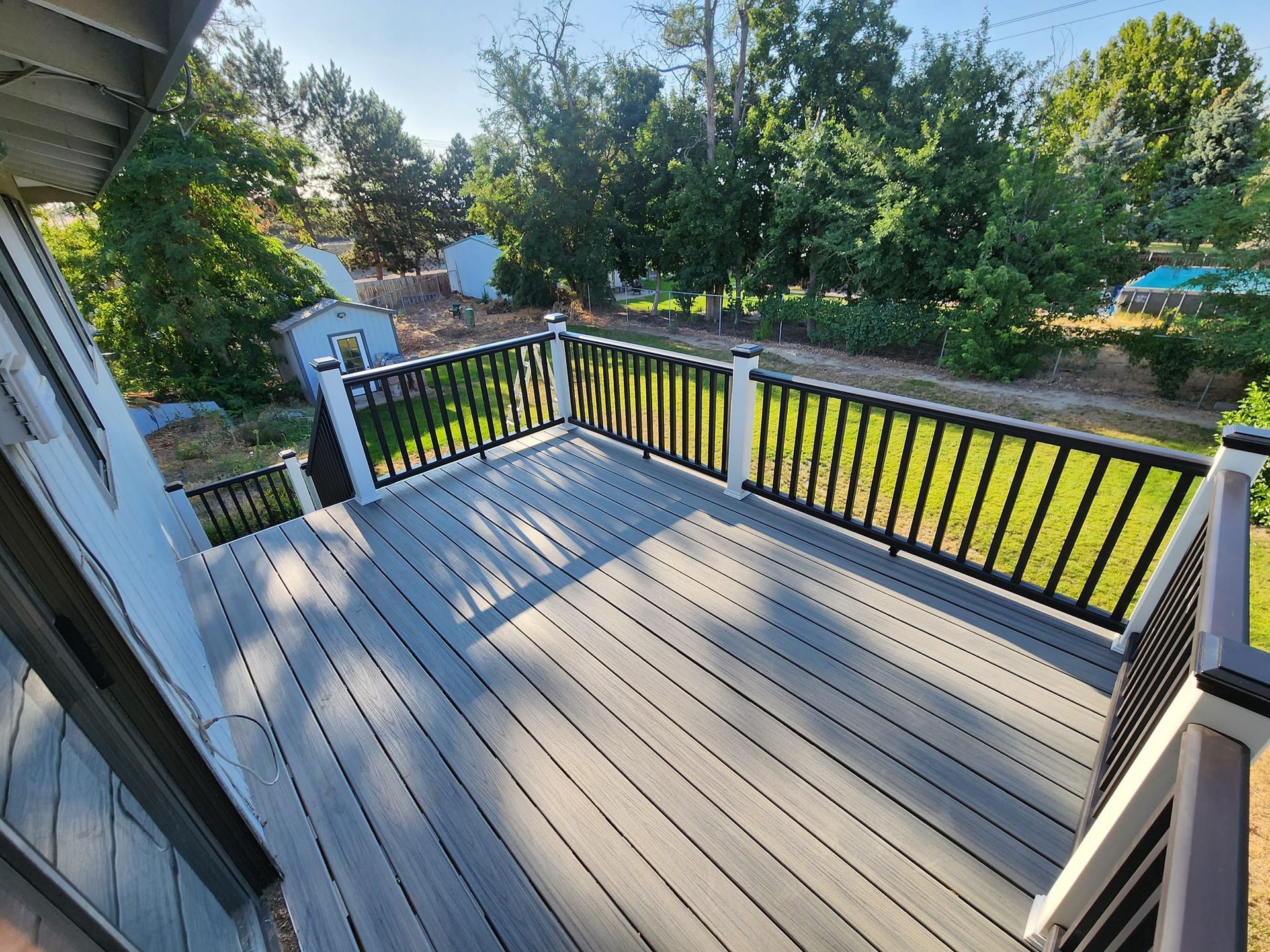 Elevated outdoor deck with gray decking, black railings, and white posts, overlooking a yard with trees.