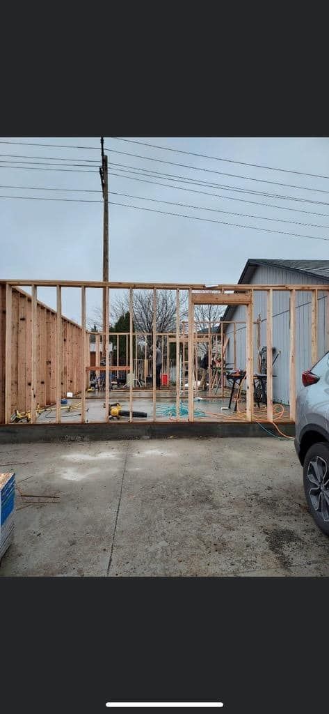 Construction of a wooden building frame, with a car parked nearby and utility lines overhead under a cloudy sky.