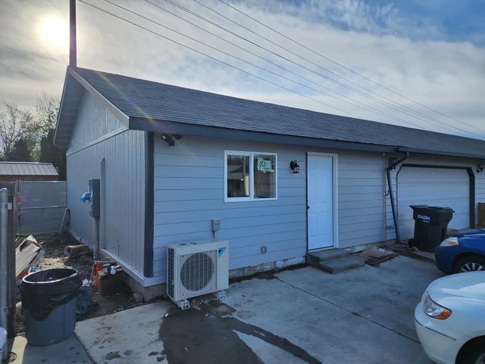 Light blue building with white door, window, air conditioner, and garage. Gray roof, overcast sky.