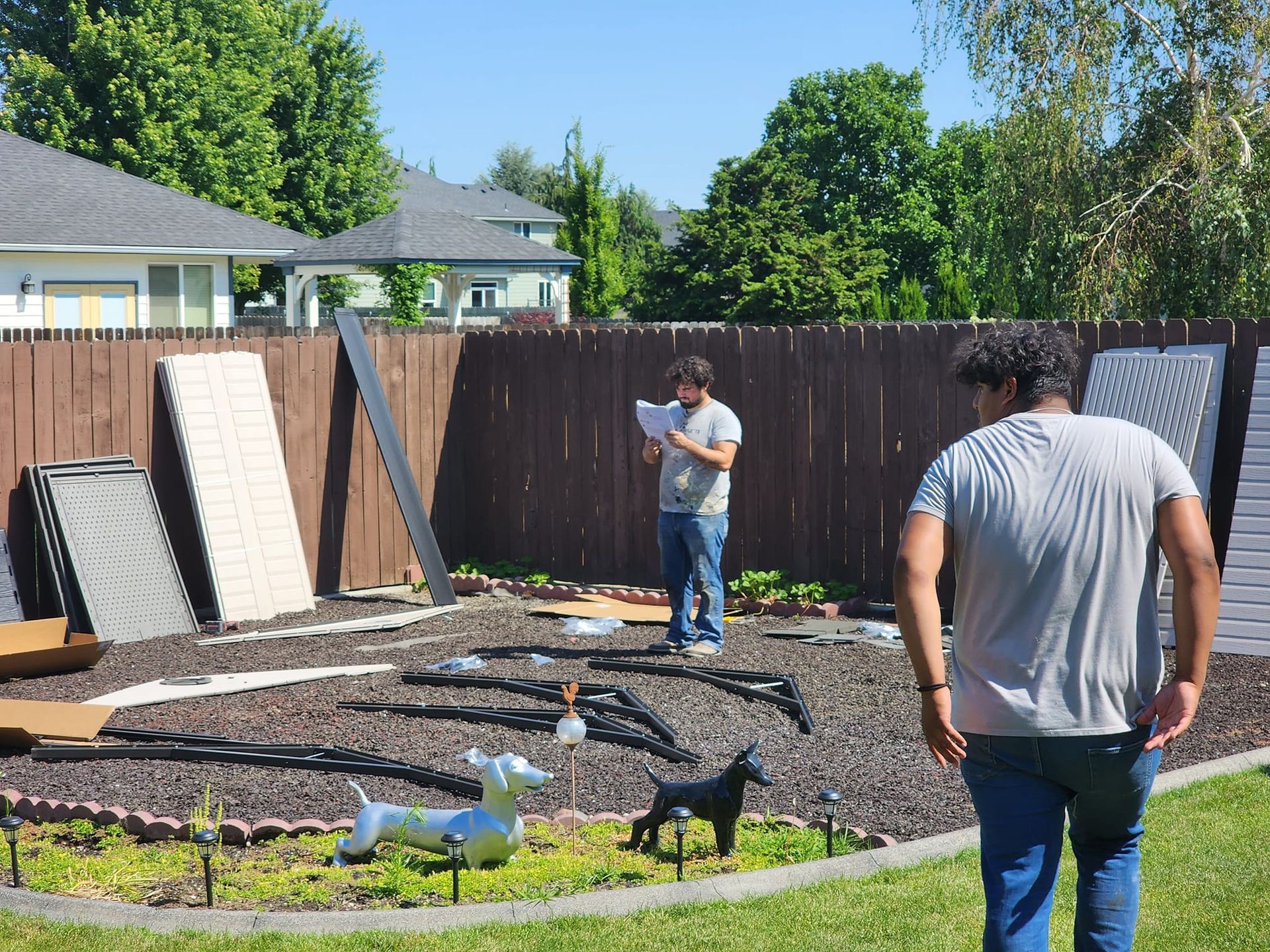 Two people assembling a structure in a backyard, surrounded by materials and garden decor. One reads instructions.