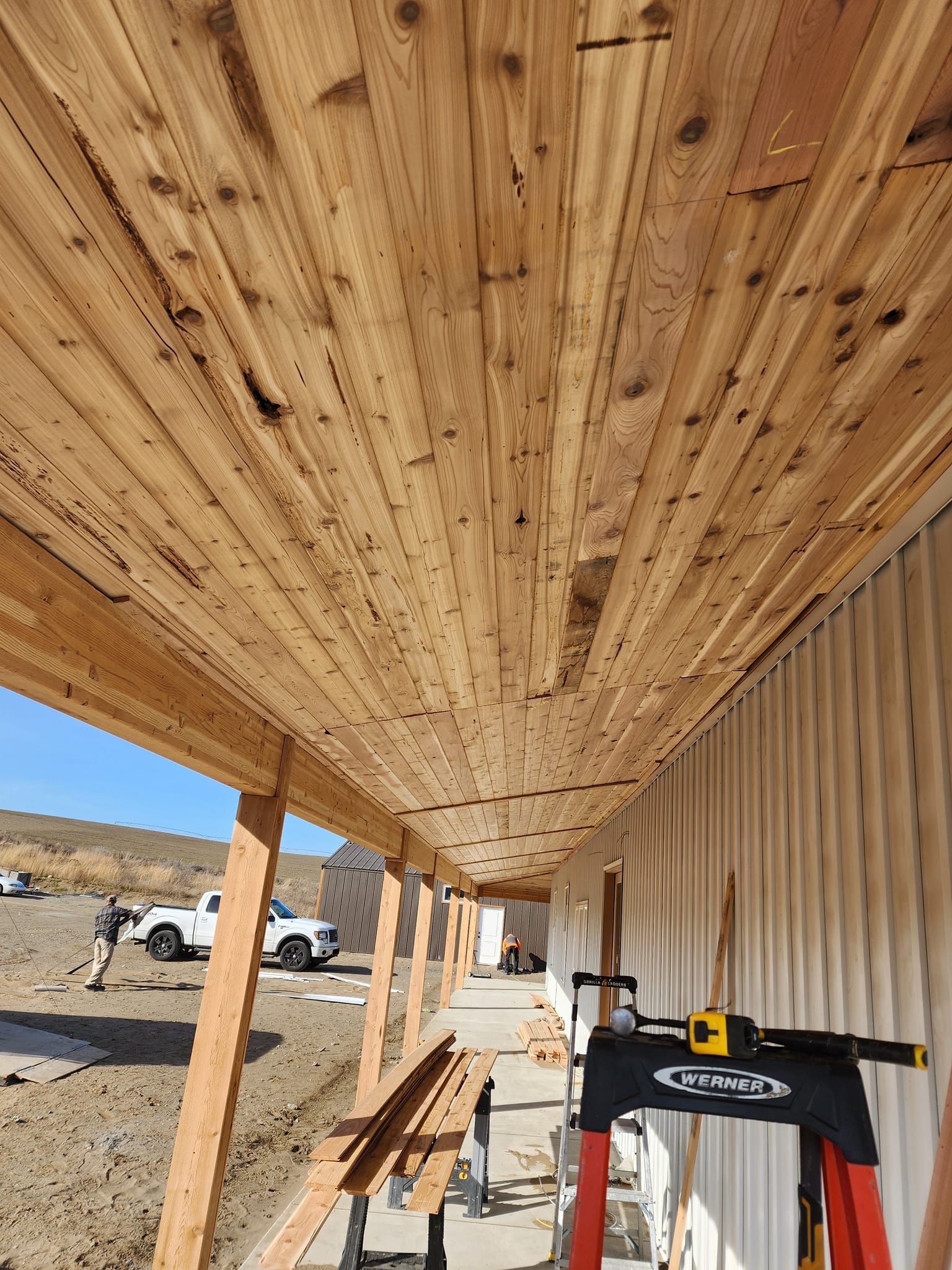 Cedar wood ceiling being installed on a porch. White siding and a sunny outdoor setting.